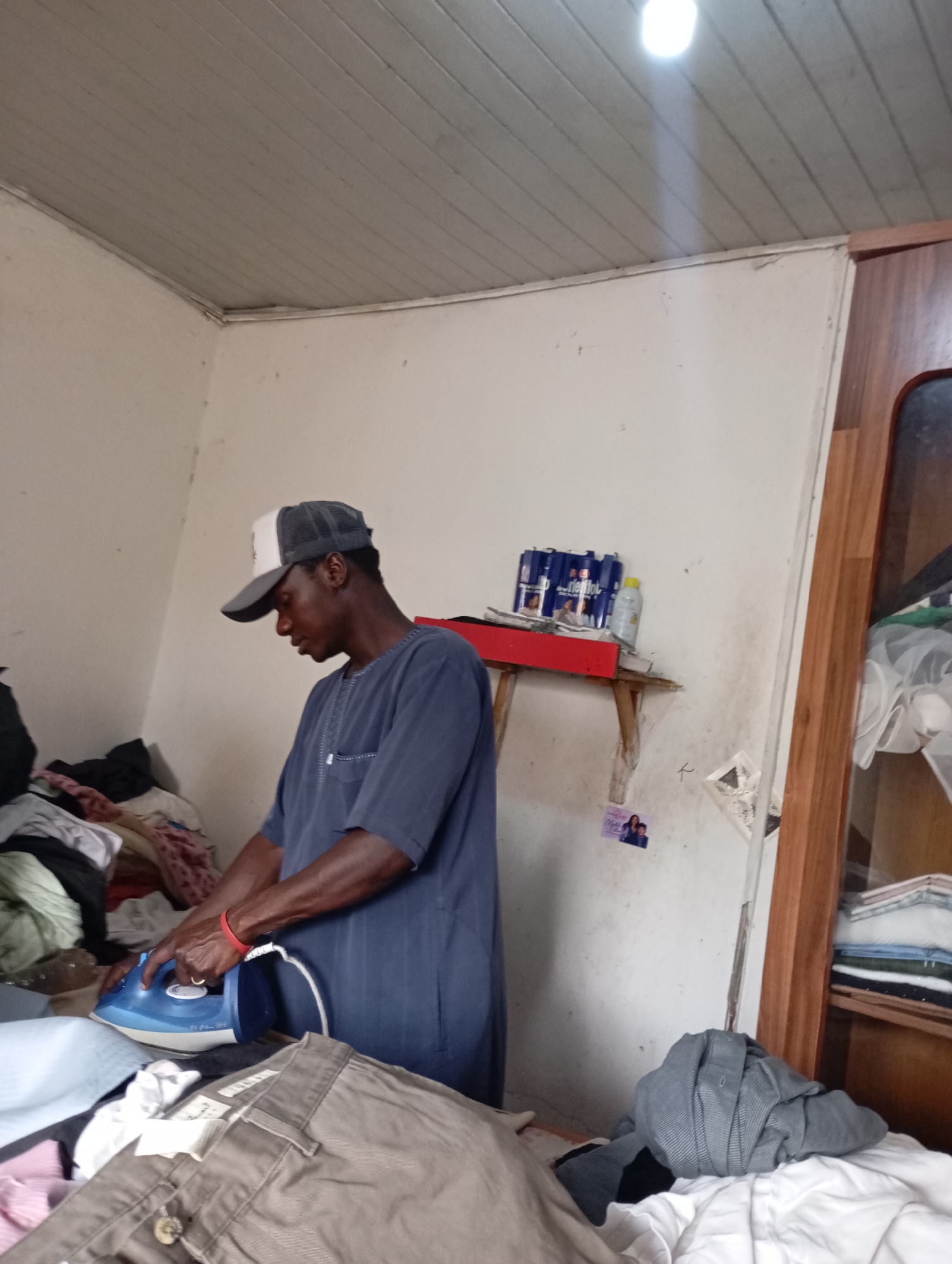 A young Black man wearing a grey and white baseball cap and a navy blue tunic is ironing clothes in a room. The room appears to be a storage or laundry area with piles of clothes on surfaces. A blue iron is being used on a brown garment in the foreground. In the background, there are shelves with canned drinks and other items. The walls are white and appear somewhat worn, and the ceiling is made of wooden planks. The scene suggests a domestic or service-oriented environment, possibly in a residential area within Jos, Nigeria.
