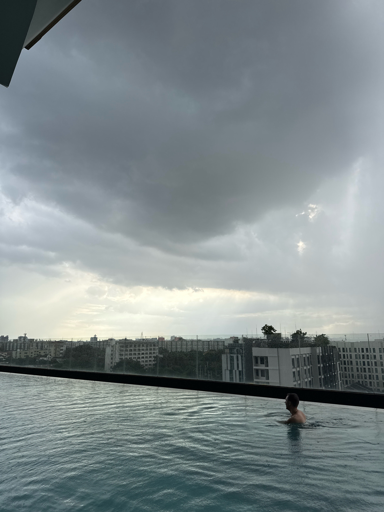 A person swims in an infinity pool on a cloudy day. The pool overlooks a cityscape, with buildings of various heights and architectural styles. The sky is filled with dark, heavy clouds, suggesting an impending or ongoing storm. The water in the pool is slightly disturbed, indicating light rain. The scene appears to be from a high vantage point, possibly a rooftop or a tall building. Based on the architectural style of the buildings and the tropical vegetation, it is plausible this scene is in Samrong Nuea Subdistrict Municipality, Thailand.