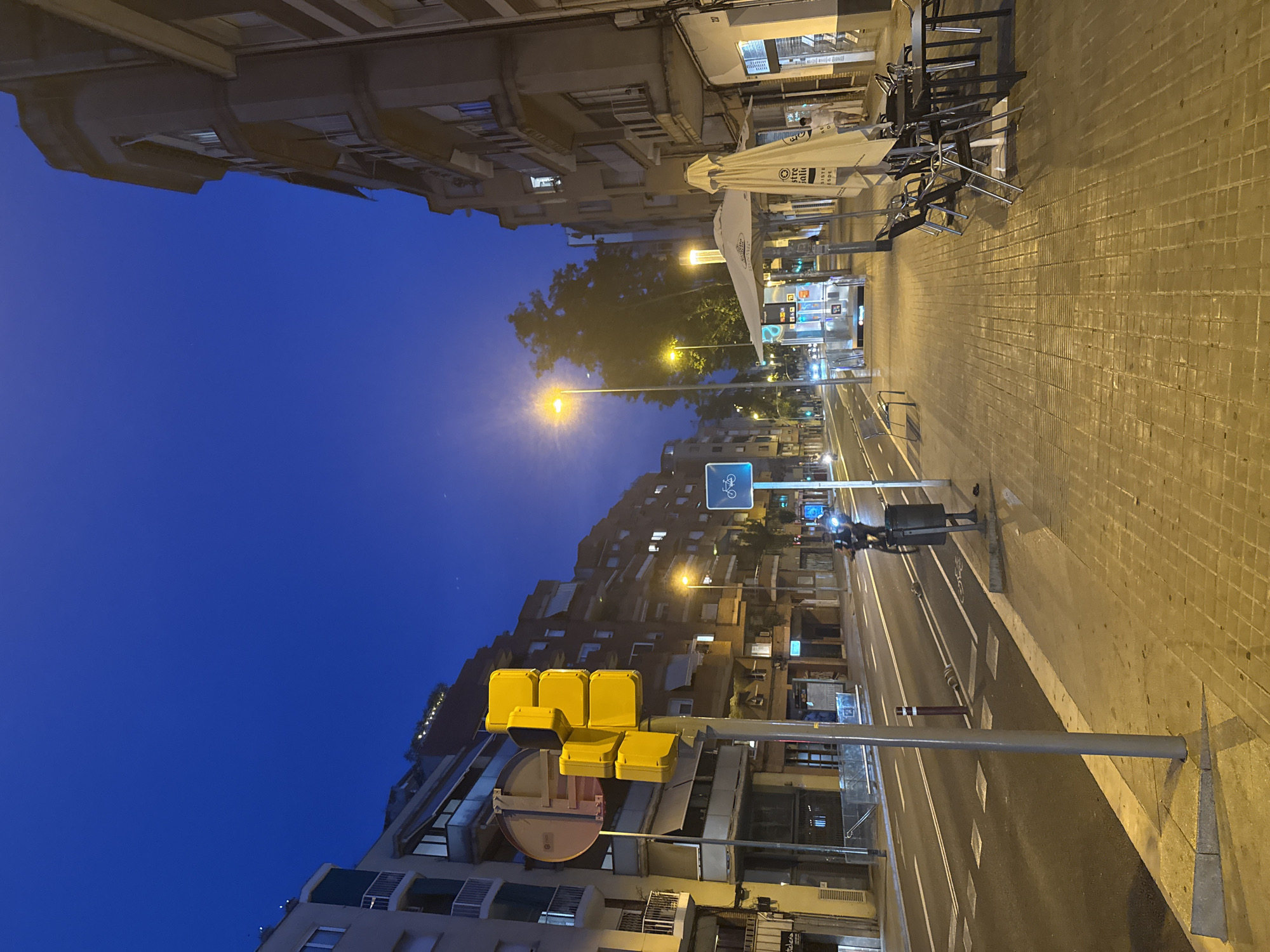 The image depicts a street scene at dusk or early night in Barcelona, Spain. The sky is a deep, clear blue. On the left side of the street, a traffic light with several yellow lights is prominently displayed. Adjacent to the traffic light, a circular sign with a red border and yellow interior is visible. Buildings line both sides of the street, with multiple stories and balconies. The street itself appears to be a multi-lane road with white lane markings.
On the right side of the street, a sidewalk is paved with textured bricks. Several outdoor tables and chairs are set up, some with umbrellas. A small structure, possibly a kiosk or bus stop, is also present. Streetlights illuminate the sidewalk and parts of the street. A blue rectangular sign with a bicycle symbol is visible on a pole.
A person on a bicycle with a headlight is riding in the street. Another person is walking on the sidewalk. A trash can is located near the curb. The overall environment suggests an urban setting.