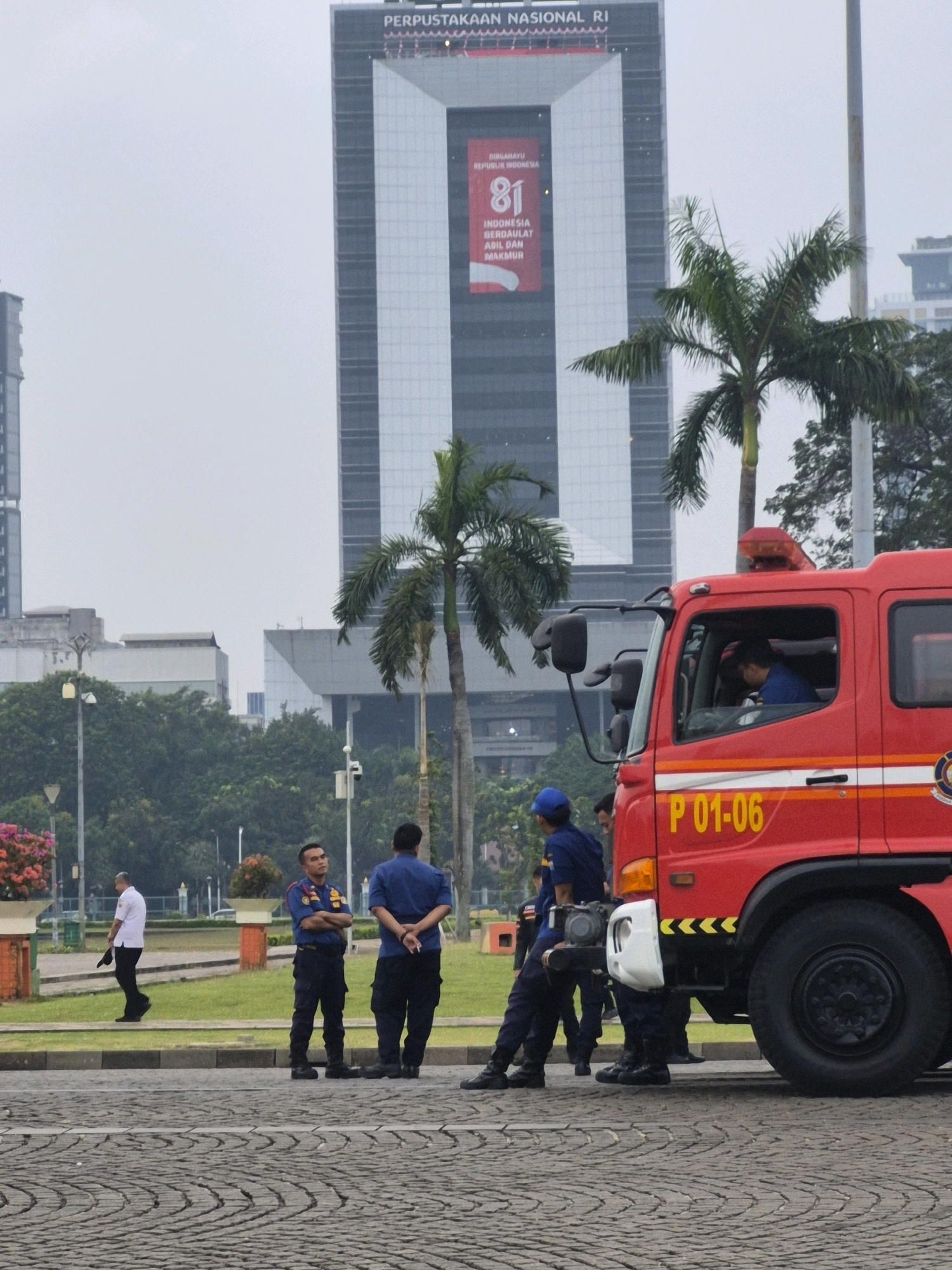 In Central Jakarta, Indonesia, a fire truck with the license plate "P 01-06" is parked on a cobblestone street. The truck is red with white and orange stripes and has a flashing light on its roof. A person in a blue uniform, likely a firefighter, is visible in the driver's seat. To the left of the fire truck, several individuals in uniforms are standing on a grassy area. One man in a white shirt and black pants is walking away from the camera, carrying a hat. Two other men in blue uniforms are standing with their arms crossed or behind their backs, facing the fire truck. In the background, a tall modern building with a large banner displaying "81" is visible, along with palm trees and other greenery, suggesting an urban environment. The sky is overcast.
