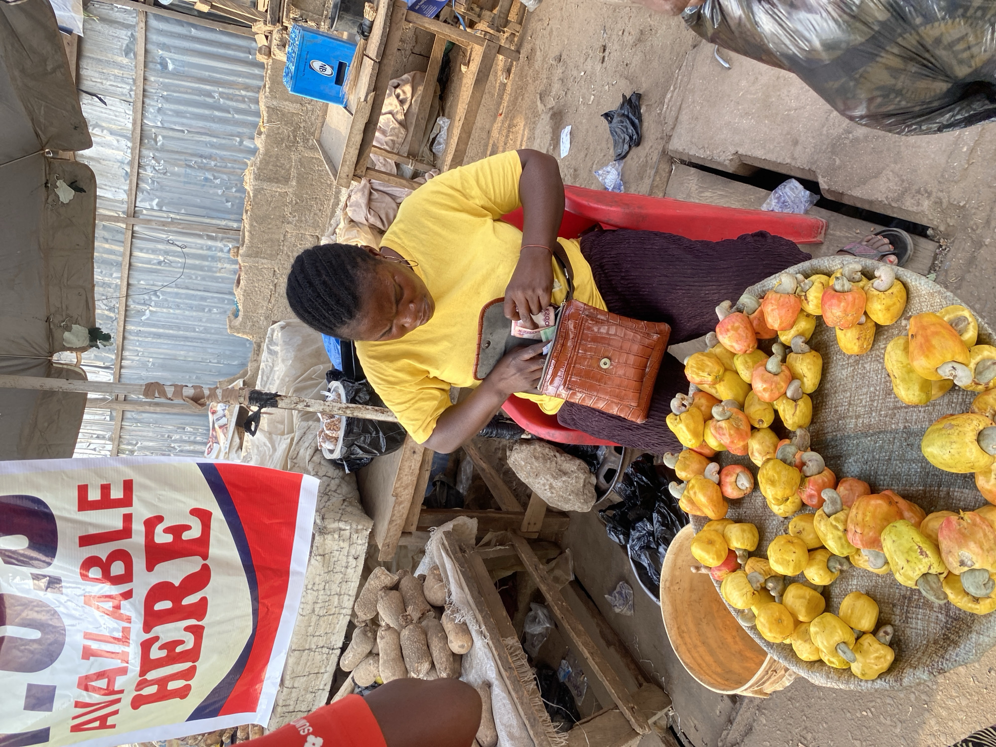 The image depicts an outdoor market stall scene in Sabon Tasha, Nigeria. A woman, wearing a yellow short-sleeved t-shirt and dark patterned skirt, is seated on a red plastic chair. She is actively handling Nigerian Naira banknotes, placing them into or retrieving them from a brown, textured, leather-like purse or wallet that rests on her lap. Her hair is styled in dark braids.
In the immediate foreground, a large, round, shallow basket contains a prominent display of numerous ripe cashew apples, predominantly yellow with some exhibiting orange and reddish hues, each with an attached cashew nut. To the left, a portion of another individual, identifiable by a red short-sleeved shirt with a white logo, is visible.
Behind the seated woman, to her left, a raised wooden table displays a pile of brown yams. Above this area, a white banner with red text prominently reads "AVAILABLE HERE." The market stall is covered by a grey canvas or tarp awning supported by metal poles. Further in the background, a rough concrete block wall is visible, topped by corrugated metal sheeting forming a fence or structure. Various wooden tables and crates, including a blue plastic crate with "NB" branding, are scattered around the scene, alongside black plastic bags and discarded refuse on the dirt ground. The setting indicates a retail transaction environment under natural daylight.