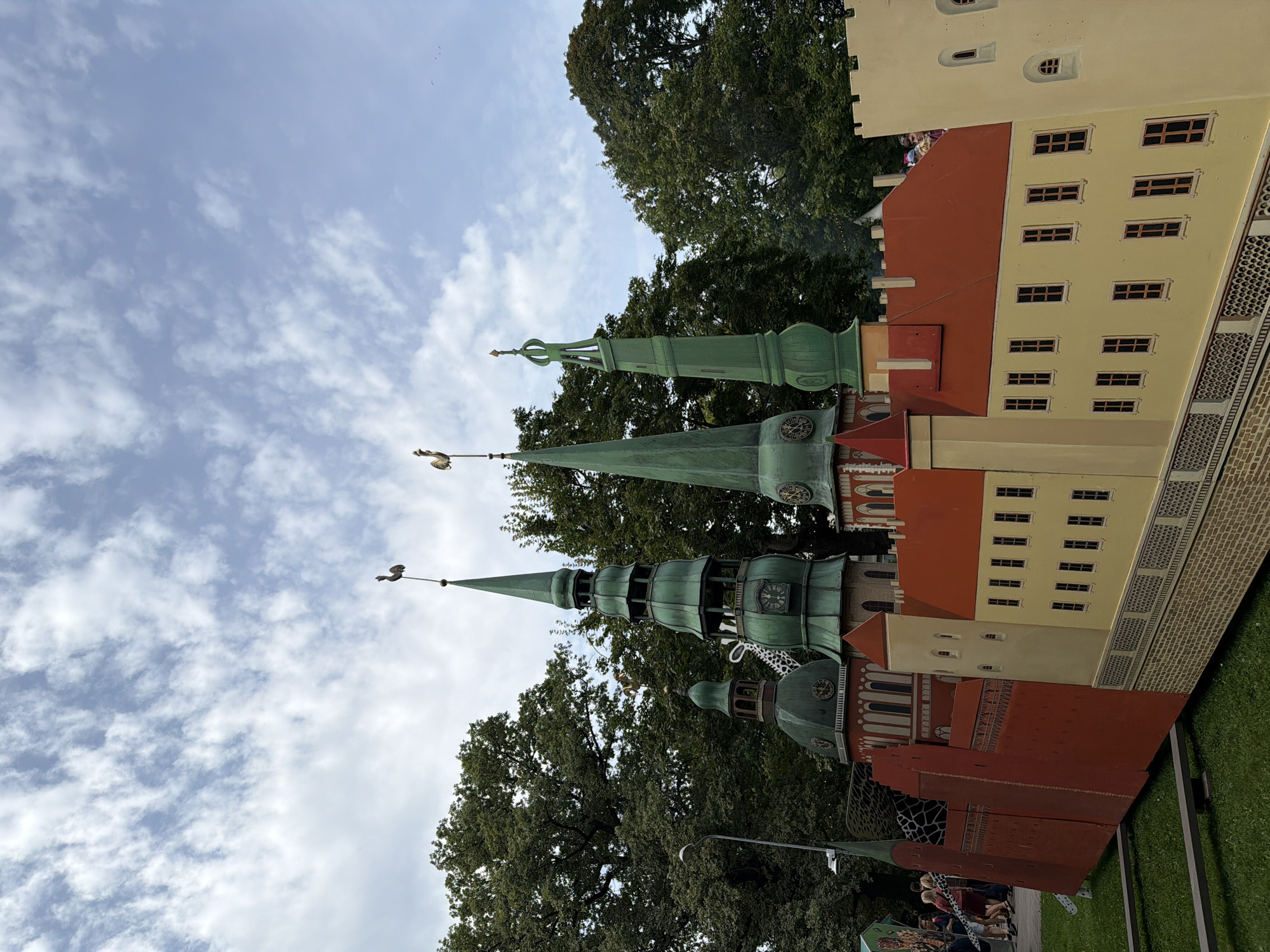 The image shows a model of Riga Castle, located in Riga, Latvia. The model features the castle's distinctive red walls, yellow buildings, and green spires topped with weather vanes. The sky above is partly cloudy, with fluffy white clouds against a blue background. Trees surround the model, providing a natural backdrop. In the foreground, there is green grass and a metal support structure for the model. A few people can be seen in the background, suggesting this is a public display. The model is detailed and accurately represents the architectural features of the actual castle.