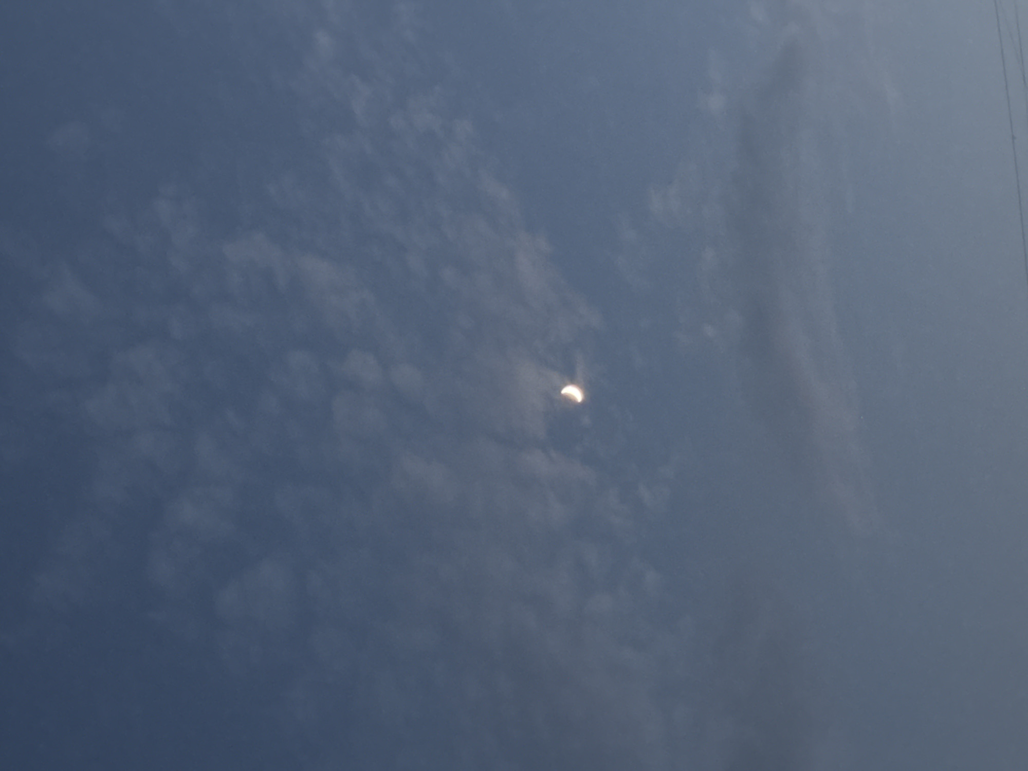 A crescent moon is visible in a hazy, pale blue sky filled with wispy clouds. The sky suggests daytime, possibly late afternoon or early morning due to the softness of the light and the presence of the moon. The overall atmosphere is serene and slightly muted. The image appears to be taken from the ground, looking upwards. There are no discernible people or man-made objects apart from a faint vertical line on the far right edge, which could be a distant utility pole or antenna. The setting is likely an outdoor location in Osh City, Kyrgyzstan, given the provided context. The weather appears to be clear but with a significant amount of haze or light cloud cover diffusing the sunlight.