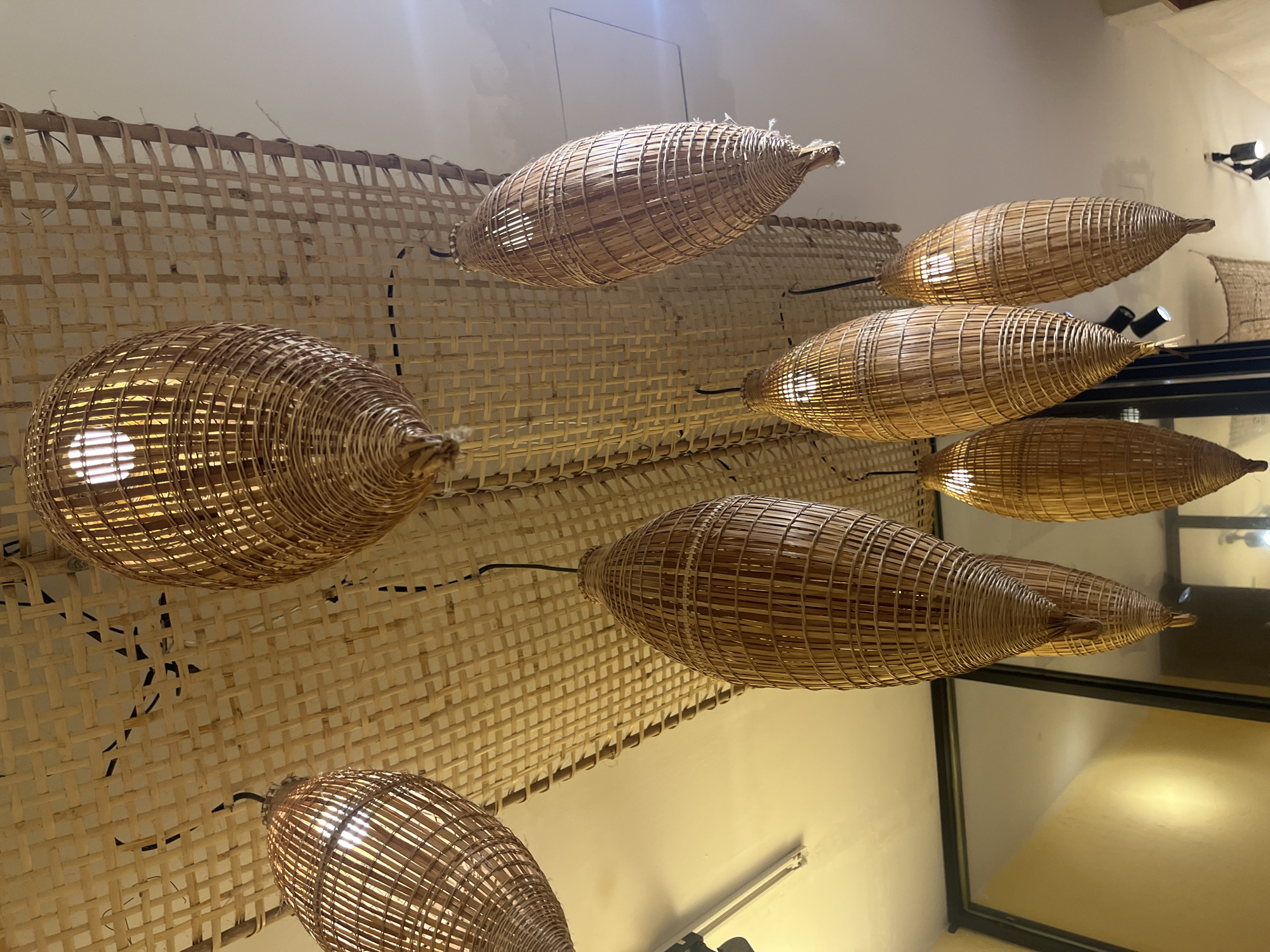 A view of a room in Nha Trang Ward, Vietnam, displays a decorative lighting scheme. Several woven basket-like light fixtures with internal bulbs cast a warm glow, suspended from a woven rattan ceiling. The room appears to be part of an establishment, suggested by the edge of a window or possibly a mirrored panel reflecting the lighting. Details like wall fixtures and furniture are visible in the reflection, contributing to the overall ambiance of the space.