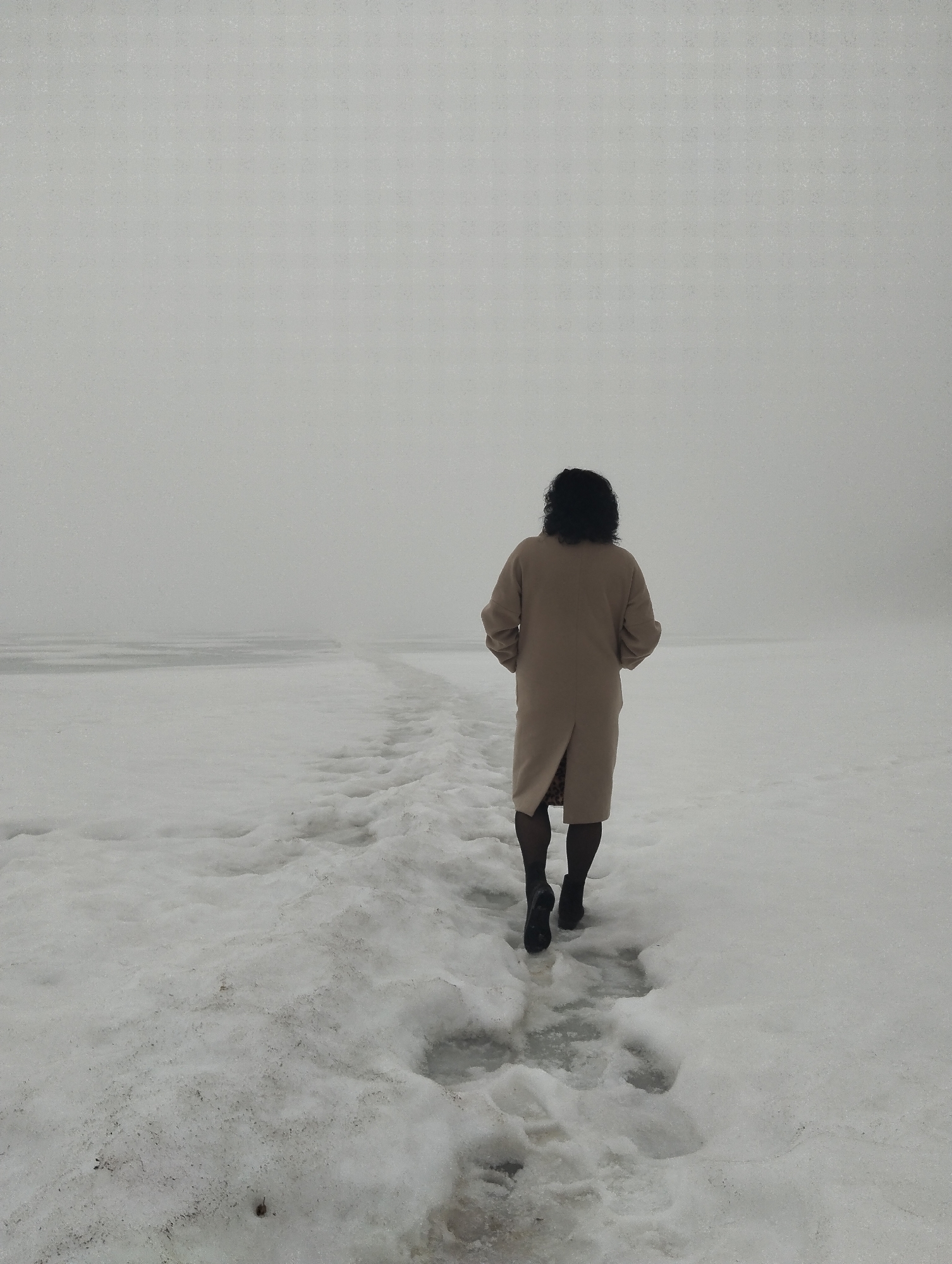 A woman in a tan coat walks away from the viewer through a snowy landscape. The snow is uneven, showing footprints and some darker patches. The background is a dense fog that obscures any distant details. The woman is wearing black tights and black boots. She is the sole human presence in the scene. The location is Votkinsk, Russia, on a frozen body of water, evident from the flat horizon line and the texture of the ground. The sky is overcast, contributing to the foggy atmosphere. The action is walking; the woman is moving forward through the snow. There is no interaction visible, only the solitary figure.