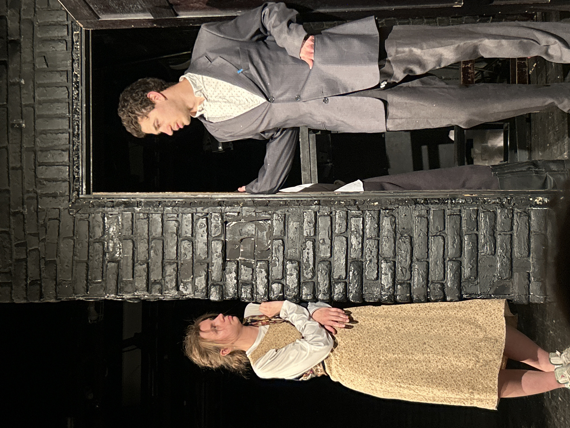 The image depicts two individuals in a theatrical setting, possibly a stage play, in an unknown city. A man in a gray suit is leaning out of a dark window frame constructed of what appears to be painted brick. Below, a woman wearing a dress and a white blouse lies in a prone position on the floor, her arms crossed over her chest. The environment appears to be dark and sparsely lit, with a focus on the two characters within their respective spaces.
