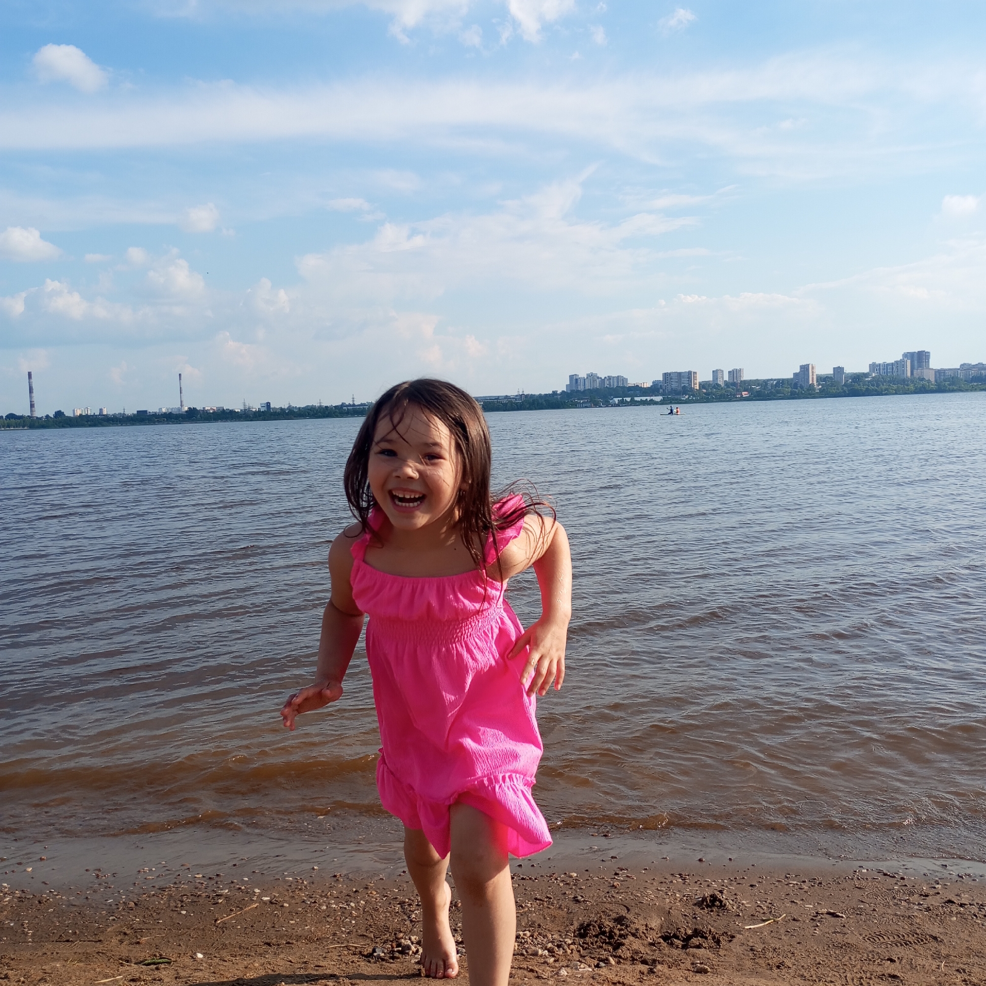 An outdoor, eye-level, full shot shows a young girl with long, dark hair, wearing a bright pink dress, standing near the edge of a body of water. She is mid-stride, with her mouth wide open in a joyful laugh, her head tilted slightly back. Her right arm is bent at the elbow, and her left arm is extended slightly forward. The water is choppy, with ripples reflecting the bright, partly cloudy blue sky. The shore is composed of sand and small pebbles. In the background, across the water, a city skyline is visible with several tall buildings and what appear to be industrial smokestacks. The location is the embankment of the Izhevsk Reservoir in Izhevsk, Russia.