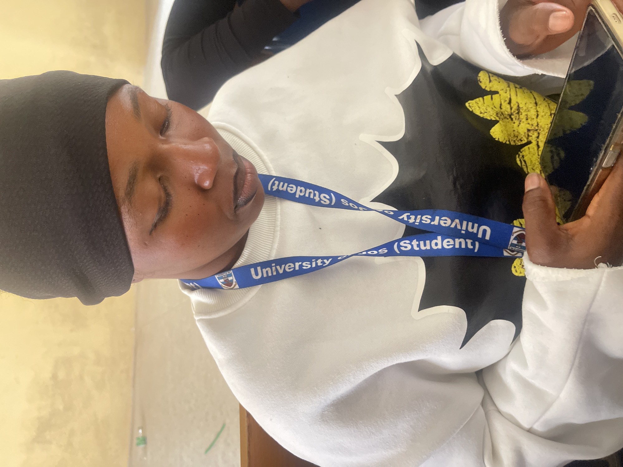 A person wearing a black headscarf and a white sweatshirt with a black and yellow graphic design is looking down at a smartphone held in their hands. The person is wearing a blue lanyard with "University of Jos (Student)" printed on it. The background appears to be an indoor setting with a yellow wall. This scene likely takes place in Jos, Nigeria, as indicated by the university lanyard.