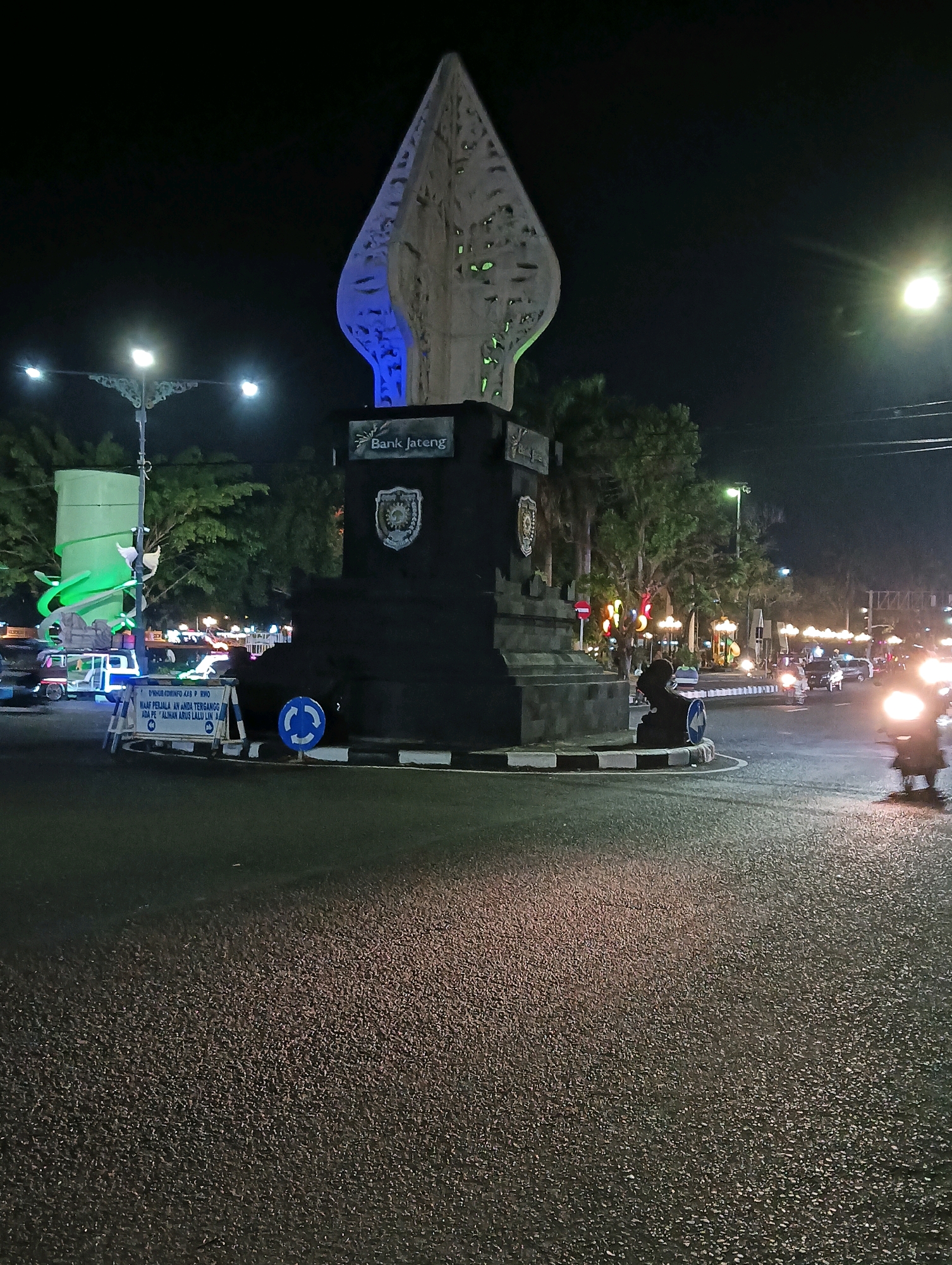 The image depicts a night scene at a roundabout in Purworejo, Indonesia, specifically the Tugu Pahlawan monument which is illuminated with blue and green lights. A prominent black pedestal supports a white, abstract monument resembling a traditional Javanese dagger (keris). The monument is adorned with a plaque that reads "Bank Jateng" and a crest. Surrounding the roundabout are trees, streetlights, and various vehicles, including cars and motorcycles. A sign on the left side of the roundabout indicates traffic restrictions. The road surface is asphalt, reflecting the ambient light. The overall atmosphere is characteristic of an urban environment at night, with artificial lighting illuminating the monument and its surroundings.
