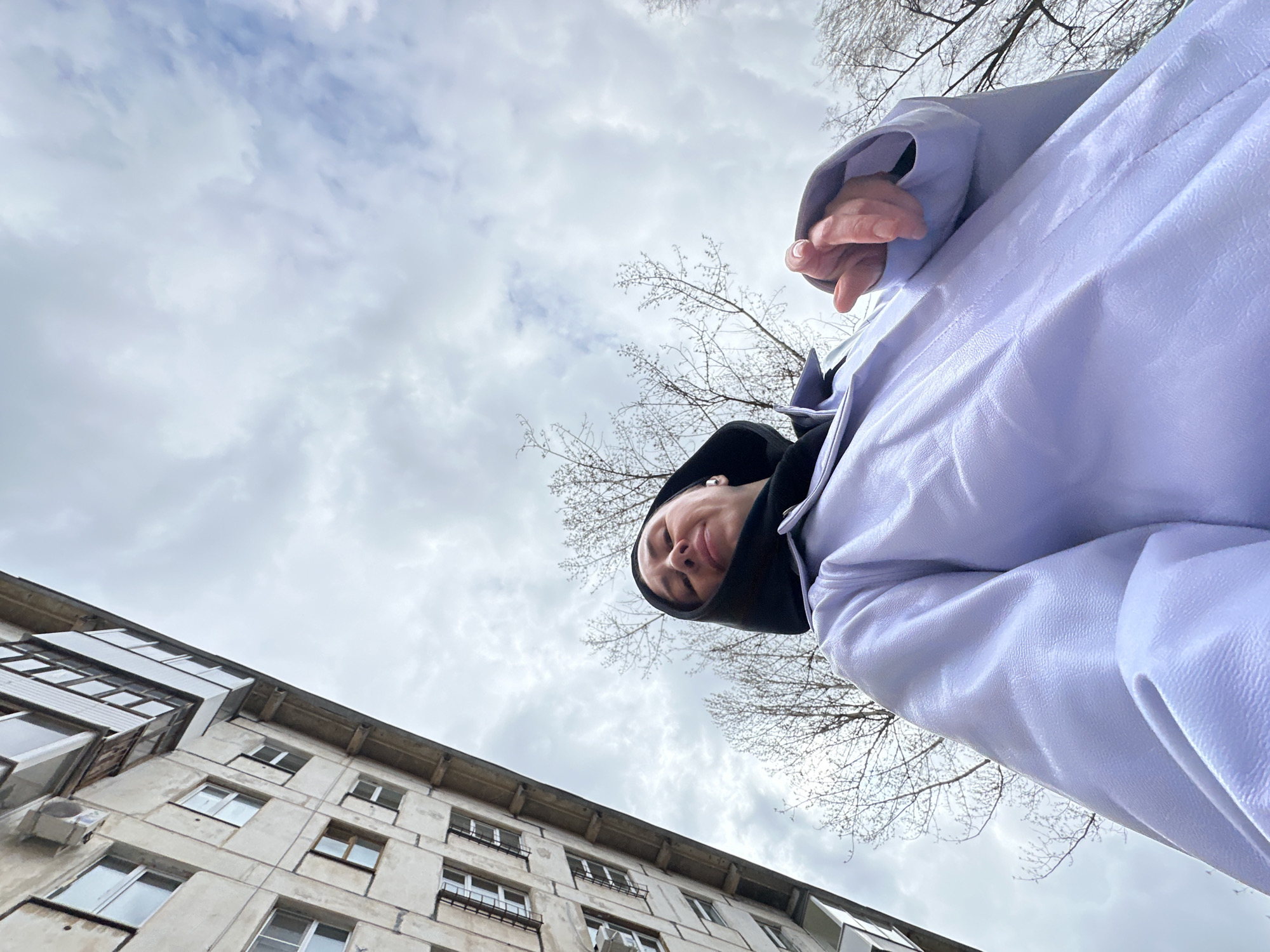 This low-angle photograph captures an individual looking down towards the camera, set against an urban backdrop under an overcast sky in Votkinsk, Russia. The subject, a person, likely female, occupies the upper right portion of the frame, gazing downwards with a subtle smile or smirk. They are wearing a light lavender-colored coat or jacket over a dark, possibly black, hooded top. An earbud is visible in their right ear, and their right hand is partially seen, emerging from the coat sleeve.
The sky is filled with varying shades of gray and white clouds, indicating an overcast daytime. On the left side, a weathered multi-story apartment building with numerous windows and balconies rises, displaying architectural characteristics common to residential structures in Russian cities. Bare tree branches, devoid of leaves, are scattered across the background on the right and center, suggesting the season is either late winter or early spring. No discernible text is visible within the image. The overall impression is a casual, possibly playful, portrait taken from a unique perspective within a typical urban setting.