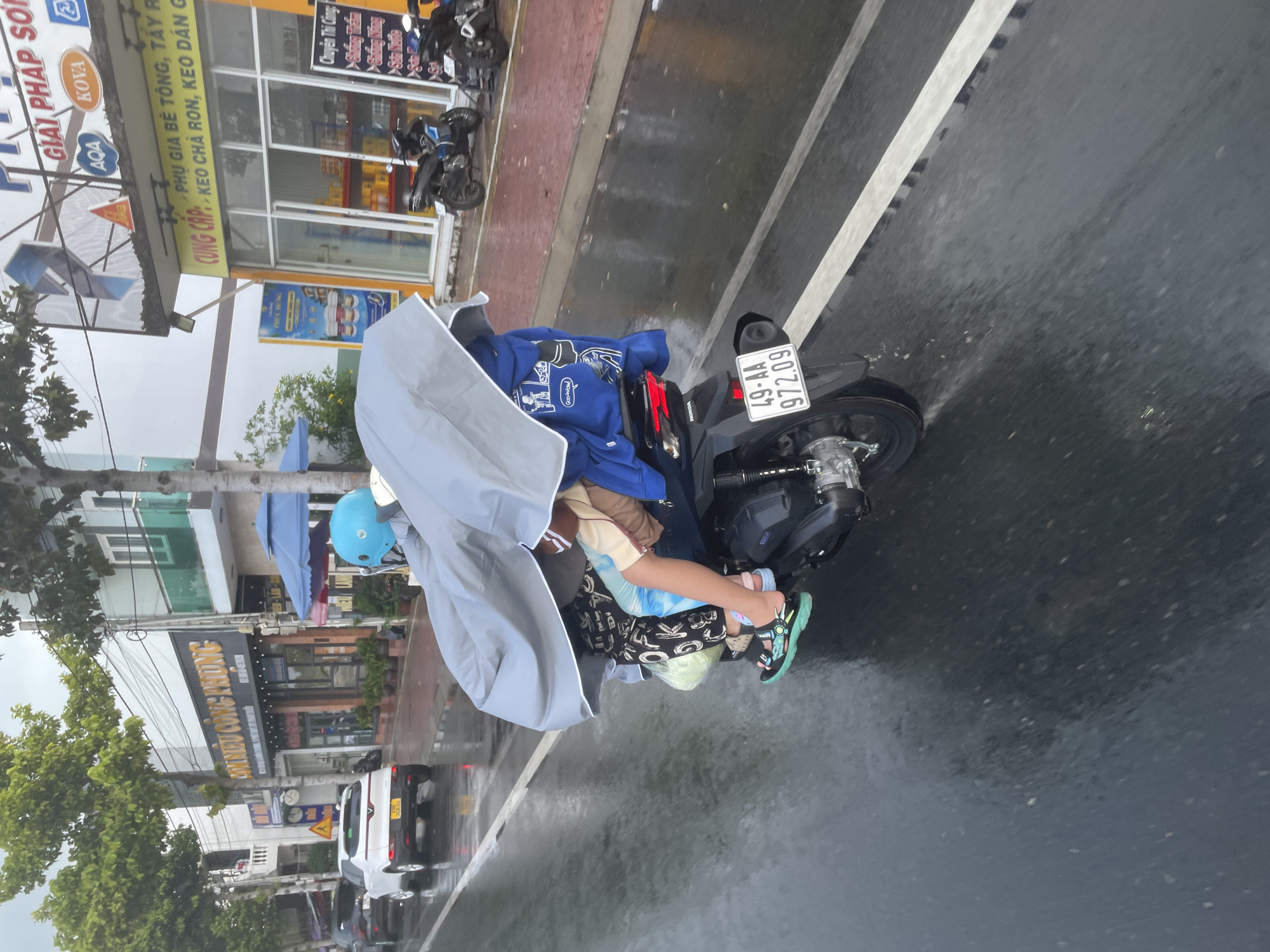 This is a close-up, eye-level, outdoor, medium shot of two people on a motorcycle during a rain shower. The motorcycle, a black Honda with a license plate reading "49-AA 972.09," is moving on a wet road. A large, gray umbrella is held over both individuals, shielding them from the rain. The person in the front, wearing a blue helmet and a light brown top, is driving. The person in the back, wearing a blue hoodie with "Goog Wow!" printed on it, is holding the umbrella and a blue backpack. The road is wet and reflective, with white lines indicating lanes. In the background, buildings line the street, displaying various signs in Vietnamese. One sign reads "GIẢI PHÁP SƠN," and another visible sign is for "CUNG CẤP KEO CHÀ RON." The scene appears to be in a commercial area, possibly Phu Thuy Ward, Vietnam, given the visible signage and the common mode of transportation. The overall atmosphere is that of a rainy day commute.
