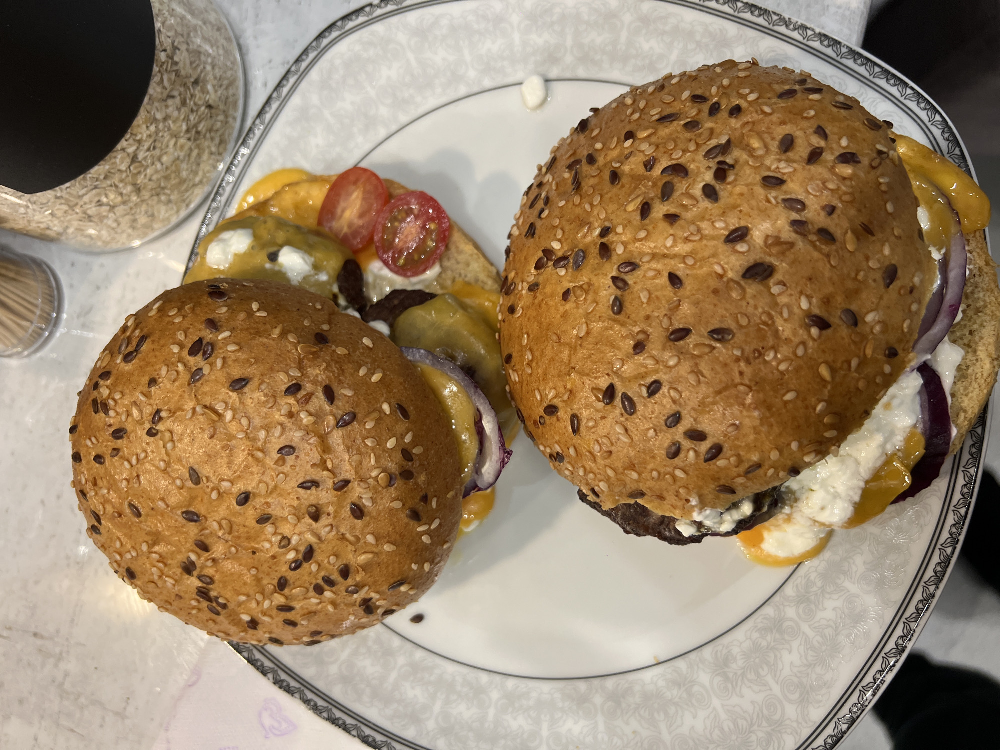 The image shows two partially assembled burgers on a white plate with a decorative gray and black border. The burger buns are sesame-seeded. One burger has the top bun removed, revealing visible ingredients including cheese, tomatoes, a meat patty, and a white topping, possibly cheese or sauce. The other burger is assembled, with visible red onion and cheese peeking out from beneath the bun. A glass container with unknown contents and a container of toothpicks are visible in the upper left corner of the frame. The location is unknown; no information present suggests Burgas, Bulgaria.