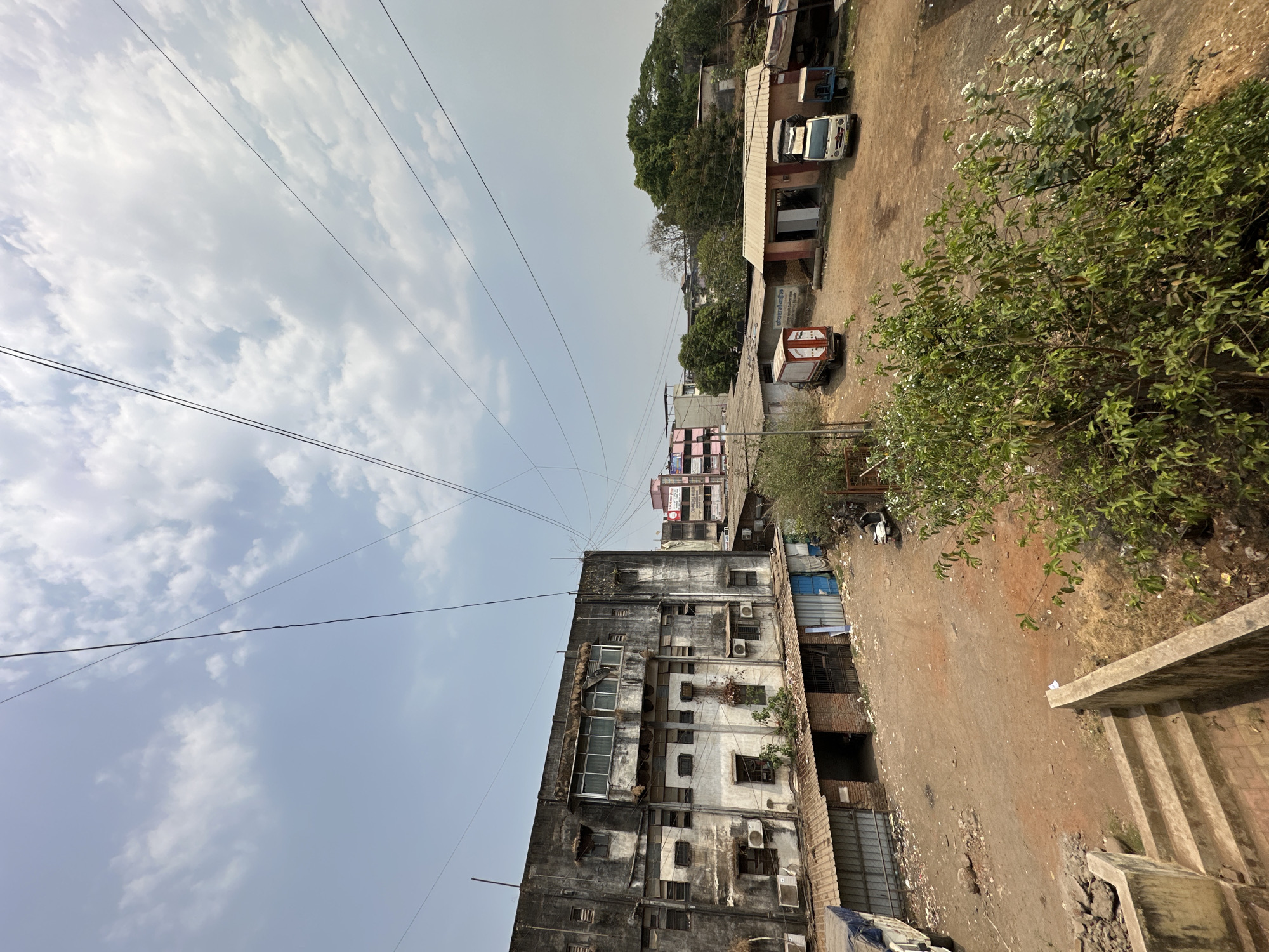 The image captures an urban street scene in Shahapur, India, under a bright, partly cloudy sky during daytime. The perspective is slightly elevated, looking down onto an unpaved, reddish-brown dirt lane or open lot that occupies the center and right side of the frame. The ground shows some unevenness and scattered debris.
On the left, a multi-story building dominates, characterized by its weathered, dark grey and white facade, numerous windows, and multiple visible air conditioning units. Utility cables and power lines crisscross the upper part of the sky, running above and around this building. Behind it, a less distinct multi-story structure with a lighter, possibly pinkish, facade can be seen.
Along the right side of the unpaved area are several smaller buildings, appearing to be single or two-story structures, varying in architectural style. A white vehicle, possibly a car or small van, is parked in front of one of these buildings. Further down, a red and white box-truck is visible; its side displays text in a local language, which appears to be "श्रीराम डेकोरेटर्स" (Shri Ram Decorators) in Marathi or Hindi, though it's partially obscured. Lush green trees and bushes are present on the far right and in the background, with a bush in the foreground featuring small white flowers. A concrete staircase descends into the bottom right corner of the image.
The sky is a pale blue with scattered white clouds, indicating a clear but not entirely cloudless day. The lighting suggests it is late morning or early afternoon, with soft shadows. No people are visible, and there is no specific event or activity taking place, depicting a typical, functional streetscape in a residential or mixed-use area.
