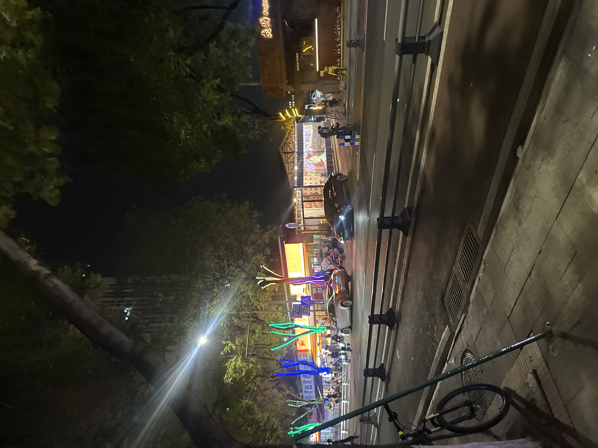 The image depicts a street scene at night in the Chongqing High-tech Zone, China. A tree with a thick trunk and branches partially obscures the view. Along the street, a row of businesses are visible, illuminated by colorful lights, including neon blue and green, and yellow. One establishment has Chinese characters above its entrance. Several vehicles are present on the road, including a silver sedan, a black sedan, and a yellow scooter. Pedestrians are seen walking on the sidewalk and near the businesses. A bicycle is parked on the left side of the image. The street appears wet, suggesting recent rain. The overall ambiance is that of a bustling commercial area at night.