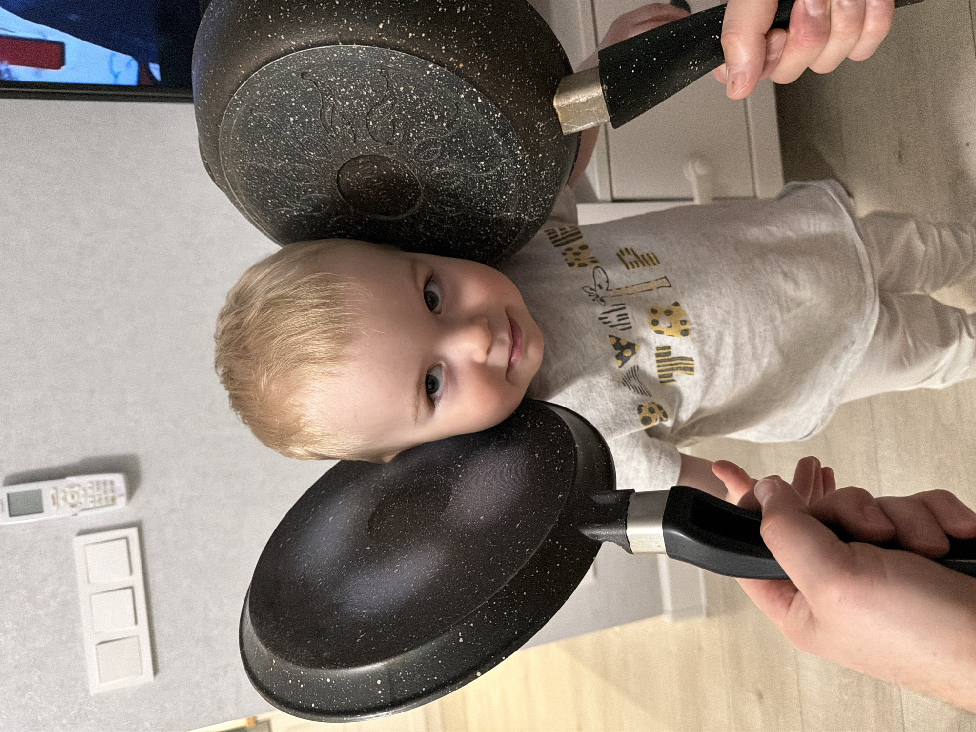 A baby is lying on their side, seemingly on a light-colored floor, with two frying pans positioned on either side of their head, apparently held by hands extending from the edges of the frame. The baby is fair-skinned with blonde hair and is wearing a light-colored long-sleeved shirt with printed designs, as well as light-colored pants. The frying pans are dark in color with speckled surfaces and black handles. In the background, there is a television mounted on the wall, a white-colored wall switch and a remote control. The setting appears to be indoors. The city is not evident in the image.
