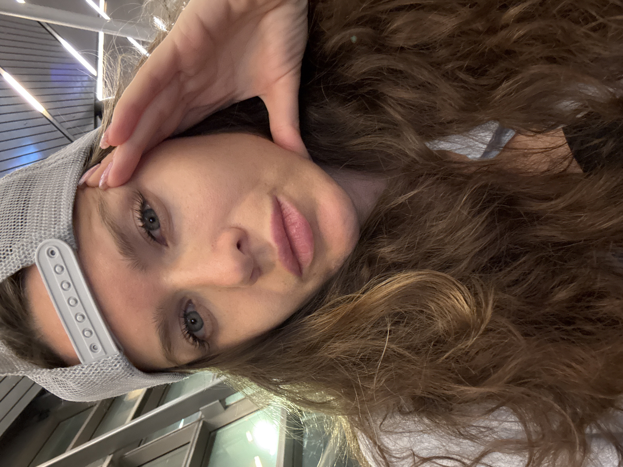 A close-up, angled selfie captures a young woman indoors, likely at Vnukovo International Airport in Russia. She looks directly at the camera with a neutral expression and slightly parted, rosy lips. Her long, wavy brown hair frames her face and cascades over her shoulders. She wears a grey mesh baseball cap backward, with its adjustable strap visible on the left. One of her hands, with light-colored fingernail polish, rests on her forehead near her eyebrow. The background features modern interior architecture, including linear light fixtures on the ceiling and striped wall or ceiling panels, indicating a large public space. No text is visible.