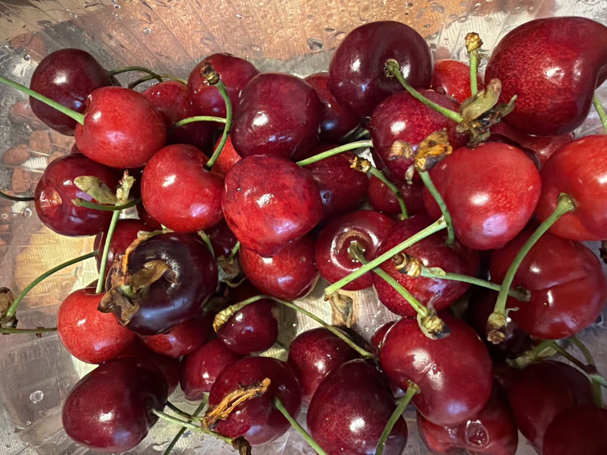 A close-up shot shows a pile of fresh cherries with green stems inside a clear plastic container. The cherries vary in color from bright red to dark red, and some have attached stems. The cherries appear to be sitting on a surface with a light brown, slightly reflective background. No people are visible. The location is not discernible from the image.