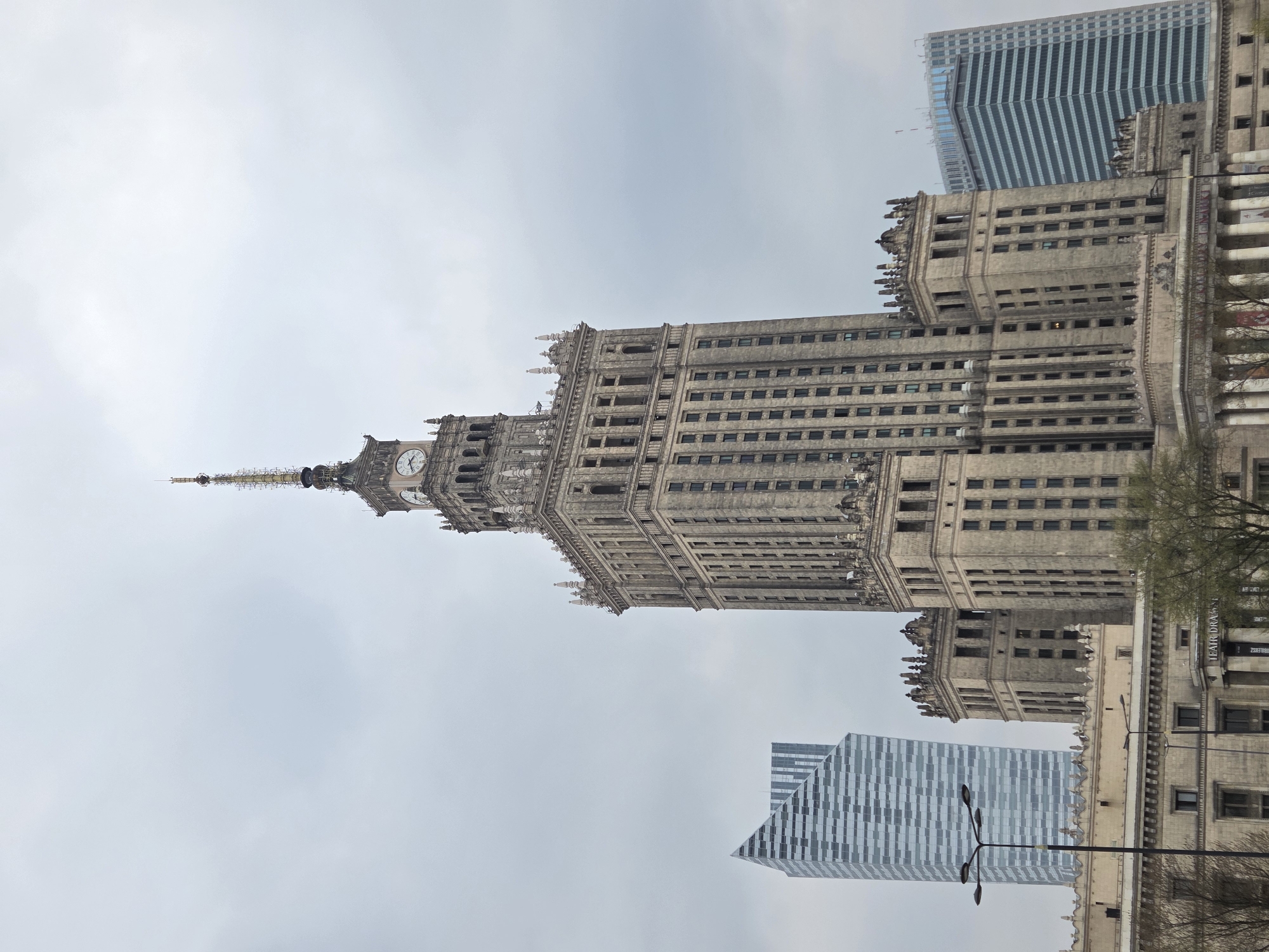 The image depicts the Palace of Culture and Science in Warsaw, Poland, surrounded by modern skyscrapers. The Palace is a tall, ornate Stalinist-era building with a prominent clock tower and spire. It is a clear day with an overcast sky. The scene includes architectural details of the Palace and the surrounding contemporary buildings.