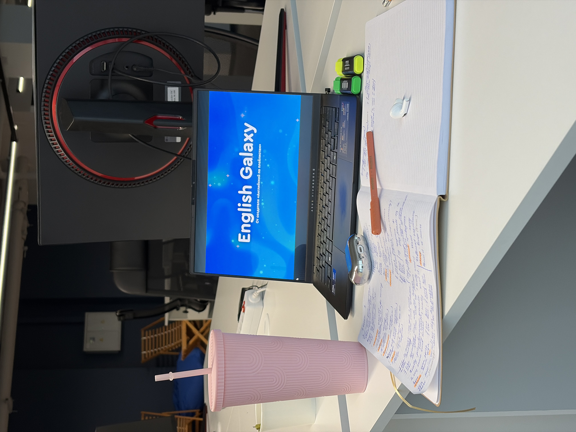 Here is a forensic description of the scene:
The scene shows a desk environment with a computer monitor and laptop positioned next to each other, along with office supplies and a beverage container. The computer monitor is black with a circular design and red accents on the back. Next to it is a laptop, open to a screen displaying the words "English Galaxy" in a large font with additional text in a smaller font below. A notebook filled with handwritten notes sits adjacent to the laptop. There are two green highlighter markers on top of the notebook. A pen and a computer mouse are also on the notebook. In front of the laptop is a pink reusable tumbler with a straw. The backdrop reveals a glimpse of the office or room's interior, with what appears to be a wooden structure and a blue object in the background. The scene is lit with bright daylight, and the objects are neatly arranged on the desk.