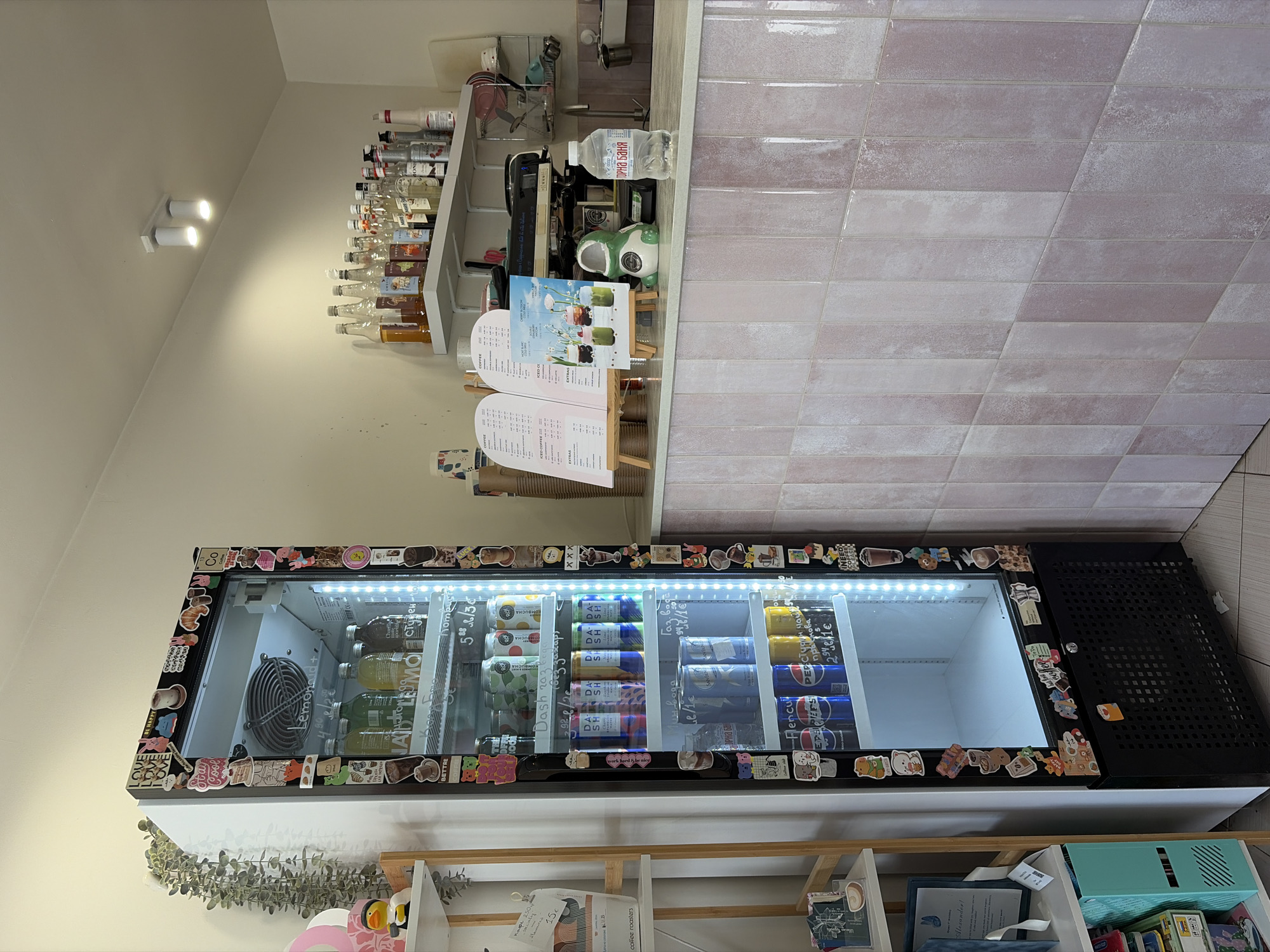 The image depicts the interior of a business, likely a cafe or similar establishment, in Burgas, Bulgaria. The walls are painted in a neutral beige color, with a section tiled in a light pink brick pattern.
A refrigerated display case dominates the lower portion of the frame. It's decorated with numerous small stickers around its frame and contains a variety of beverages, including Lemonoid, Dash drinks, and other products labeled with Cyrillic text. A shelf with storage containers and books is placed beneath the refrigerator.
Above the refrigerator, there's a counter area. A menu stand with pricing in Bulgarian currency (euro) is visible. Behind the counter is a shelf with a variety of bottles, possibly syrups or other ingredients for drinks. A water bottle labeled "Горна Баня" (Gorna Banya) sits on the counter. A coffee machine is also visible.
The lighting is provided by ceiling-mounted spotlights. Overall, the scene suggests a modern, possibly quirky, cafe setting in Burgas, Bulgaria.