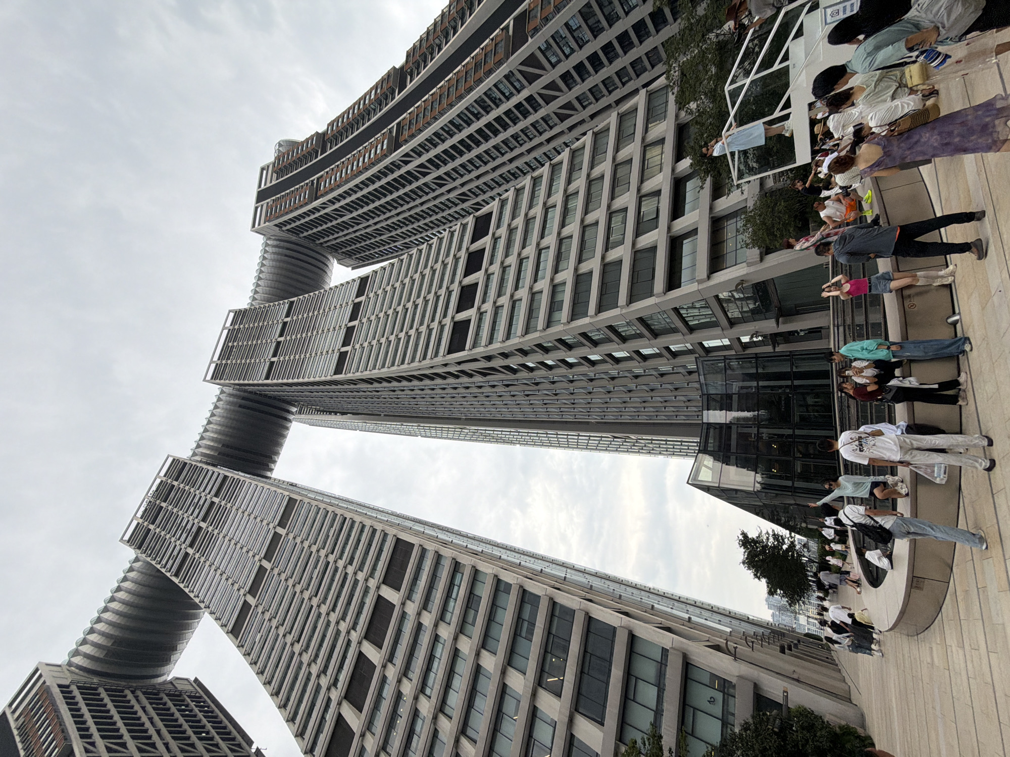 This is a wide-angle, low-angle shot of the Raffles City Chongqing complex in Yuzhong District, China, taken on an overcast day. The complex consists of eight interconnected skyscrapers, with two prominent towers connected by a massive, curved skybridge that resembles a ship's hull. The buildings are characterized by their modern, angular architecture and a facade of windows and dark paneling.
In the foreground, a plaza area with a curved fountain feature is populated by a diverse group of people. Several individuals are walking, standing, or sitting, engaged in various activities. Some are looking at the buildings, while others appear to be interacting or posing for photos. Clothing is casual and modern, with a mix of t-shirts, shorts, dresses, and jeans. The plaza itself is paved with light-colored stone tiles.
To the left of the plaza, a section of another building with a stepped, geometric facade is visible. Lush green trees are scattered throughout the scene, providing a contrast to the urban landscape. The sky is a uniform grey, suggesting cloudy weather. The overall impression is one of a bustling urban environment featuring striking contemporary architecture and public spaces.