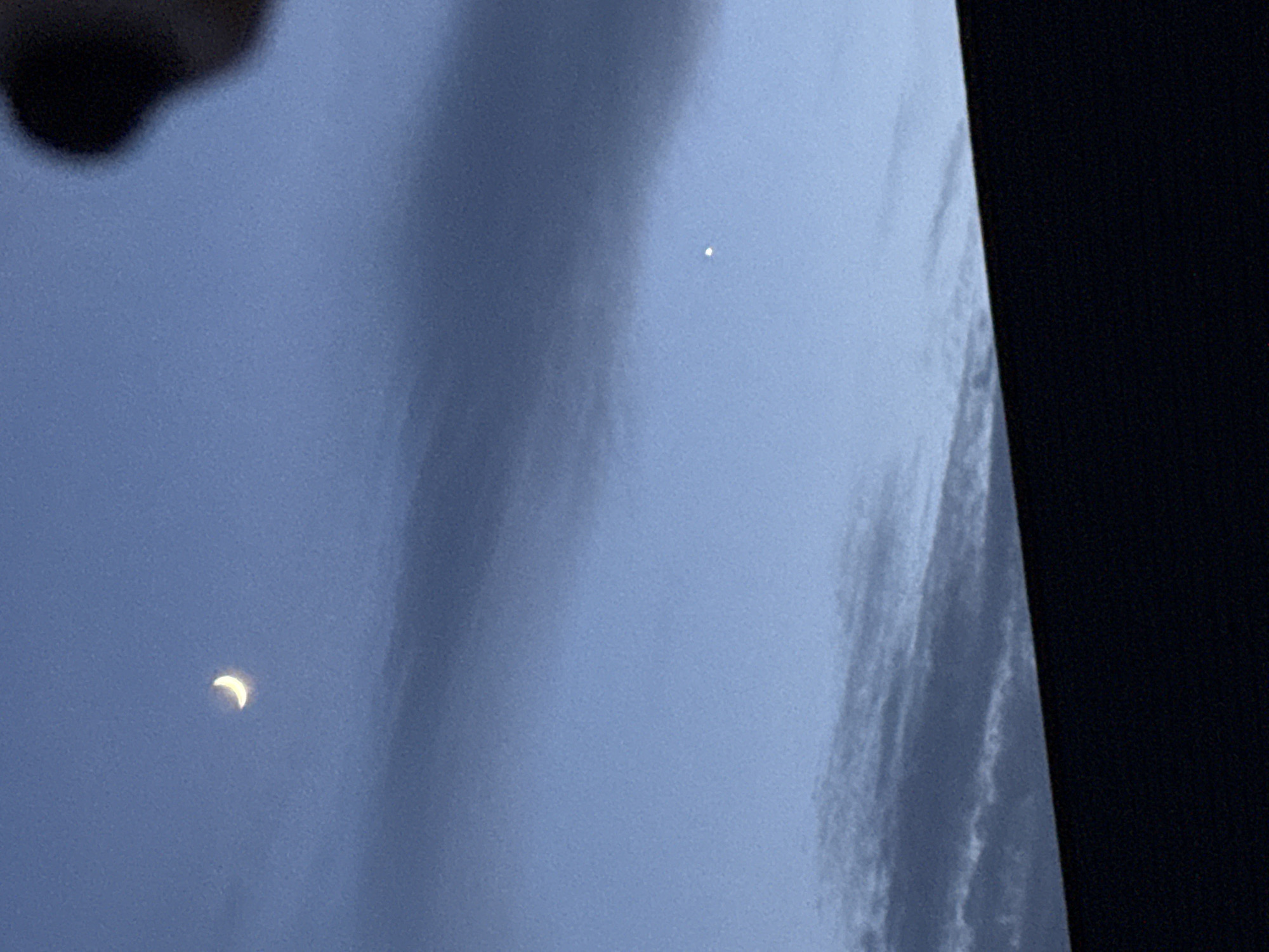 A photograph depicts a twilight sky over Great Yarmouth, United Kingdom. A thin crescent moon is visible in the lower left quadrant, and a single bright star or planet (possibly Venus) is located in the upper right quadrant. The sky is a gradient of blue, transitioning to a darker hue at the right edge of the image where a section of a dark building or object is visible. A blurred object intrudes from the upper left. A diffuse band of clouds stretches diagonally across the center of the frame. No people are visible.