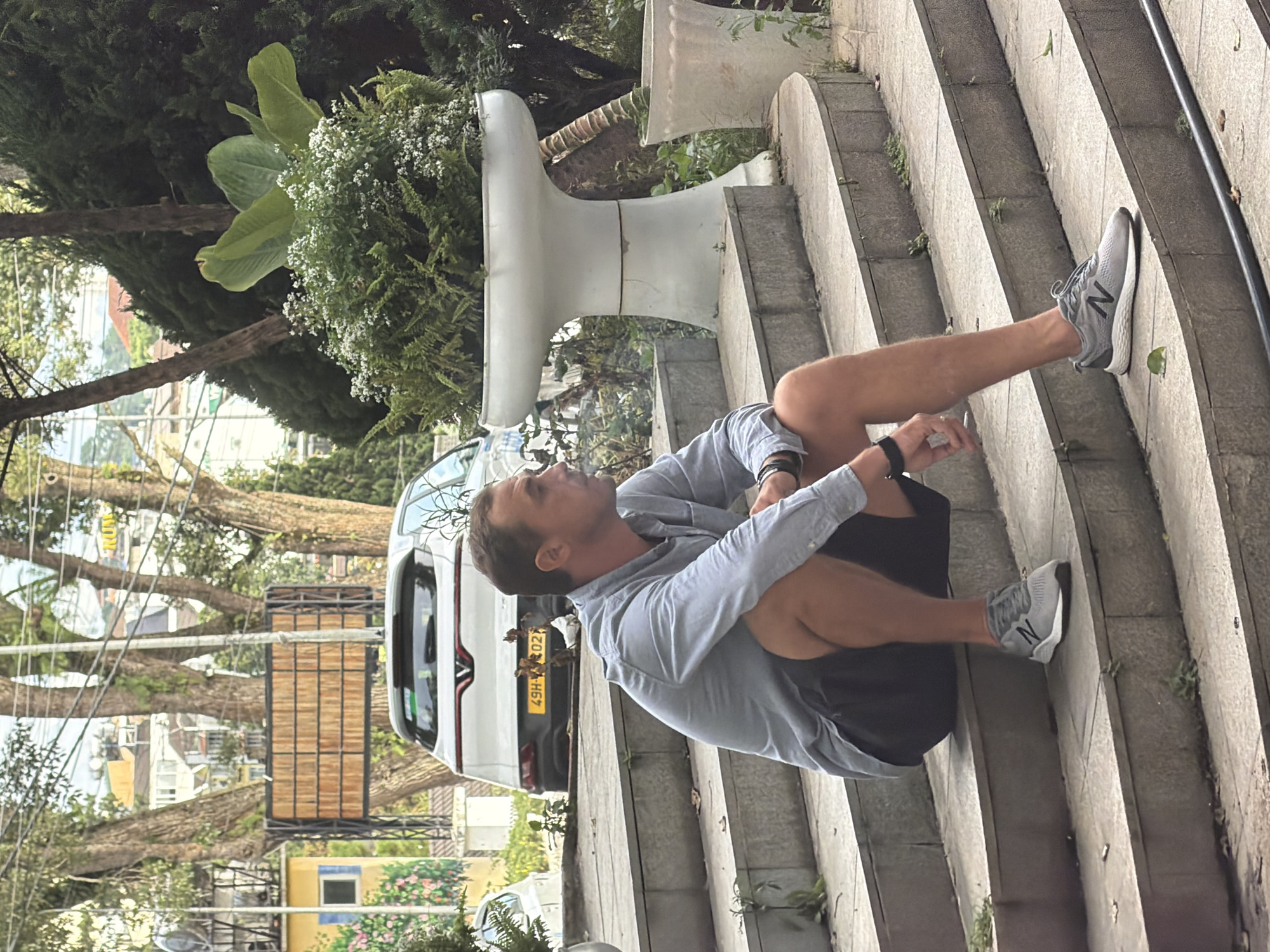 The image shows a man sitting on a set of outdoor concrete stairs. He is wearing a light blue button-down shirt and black shorts. His right leg is extended, and he is wearing a gray athletic shoe with a white sole and black laces. His left leg is bent, with his foot resting on the step below. He is looking upwards and to his left, with a thoughtful expression. A black watch is visible on his left wrist.
Behind the man, a white car with a license plate that reads "49H2-302.22" is parked. The car appears to be a hatchback. Further in the background, there are trees with lush green foliage, and hints of buildings are visible. A large, white, horn-shaped object is situated on the stairs to the man's left, possibly a decorative element or a piece of outdoor furniture. The scene appears to be outdoors, with natural light suggesting it is daytime. The presence of the car and the overall greenery suggest an urban or semi-urban environment.
The image has been rotated such that the scene appears vertical. The orientation of the subjects and objects suggests a tilt of approximately 90 degrees clockwise from the natural orientation. The location context provided is Da Lat, Vietnam.