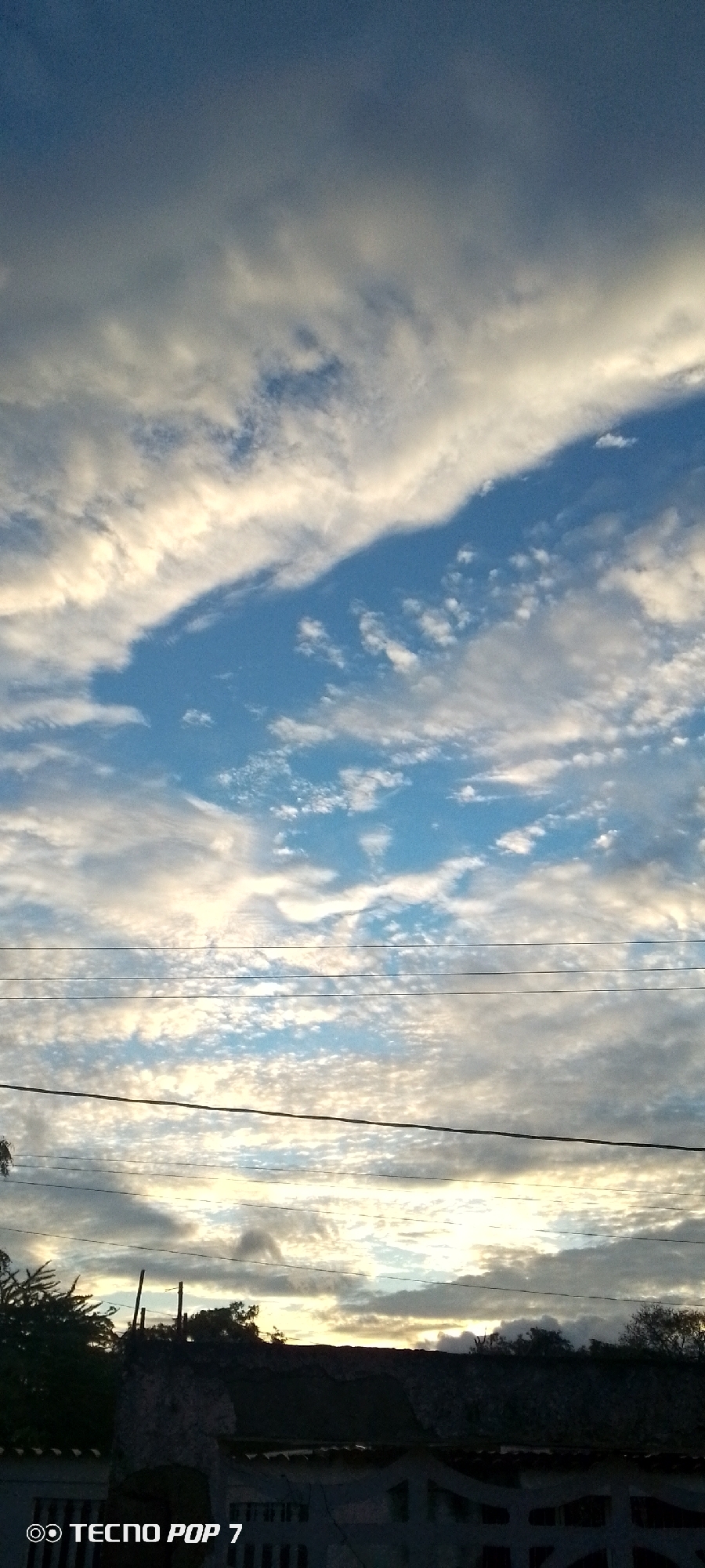 This is a vertical, low-angle outdoor shot of the sky above Altagracia, Venezuela. The sky is filled with a mix of blue sky and clouds, with some clouds appearing white and bright due to sunlight, while others are darker and cast shadows. The clouds are textured and appear to be moving. Three horizontal power lines stretch across the middle of the frame. Below the power lines, the silhouette of rooftops, trees, and some structures is visible against the brighter sky. A watermark at the bottom left reads "TECNO POP 7".