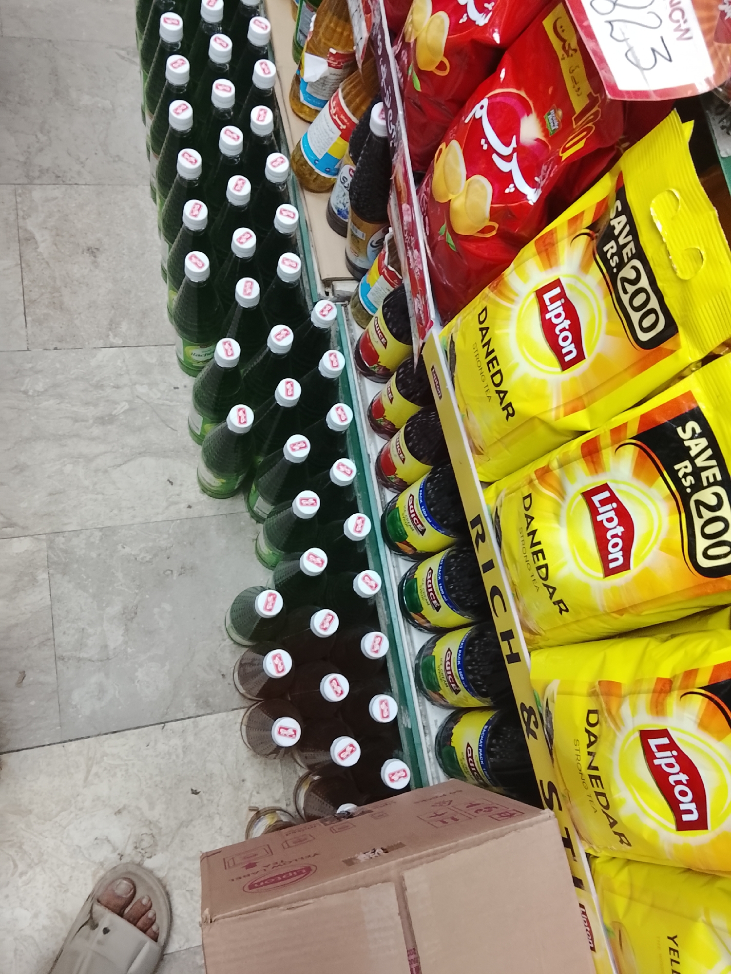 The image shows a section of a store aisle. Several rows of green and brown bottled drinks with white caps are visible on a shelf. To the right, there are stacked packages of Lipton "Danedar" strong tea in bright yellow packaging with "SAVE Rs. 200" printed on them. In front of the tea, there are red bags of chips. A cardboard box is on the floor, partially obscuring the bottles. In the lower left corner, a person's foot wearing a white sandal is visible. The location may be Gulgasht Colony, Pakistan, based on the "Rs." currency abbreviation on the tea packaging, often used to denote Pakistani Rupees.
