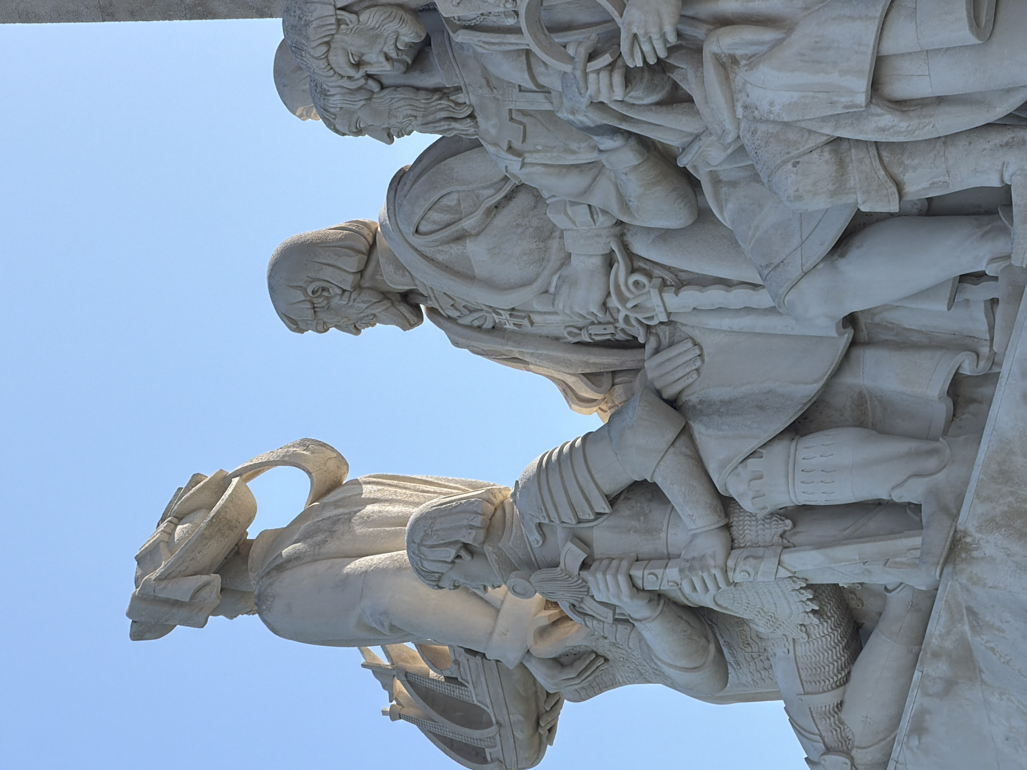 The image features a low-angle shot of a stone sculpture depicting several figures against a clear blue sky. The sculpture appears to be made of a light-colored stone, possibly marble or limestone, and is weathered, showing some signs of age and dirt. The figures are depicted in a dynamic, perhaps historical or mythological, scene.
In the foreground, a figure is seen from the chest up, wearing armor and holding a sword. This figure is positioned below another group of figures. The upper group consists of several individuals, with one prominently displayed figure in the center wearing a cloak and holding a scroll or a long, thin object. Other figures are partially visible, suggesting a group composition. The carving style is detailed, with folds in clothing and musculature clearly rendered.
The figures' expressions are difficult to discern due to the distance and the nature of stone sculpture, but their postures suggest a moment of action or contemplation. The lighting indicates it is daytime, with bright sunlight illuminating the sculpture and casting some shadows. The sky is a uniform, cloudless blue, suggesting fair weather.
The style of dress and armor hints at a historical setting, possibly from the Age of Discovery given the location context provided (Lisbon, Portugal). While no text is explicitly visible in the image, the sculpture's subject matter and style are likely indicative of a significant historical or cultural monument within Lisbon. The overall impression is one of grandeur and historical significance, captured from a unique perspective looking upwards.