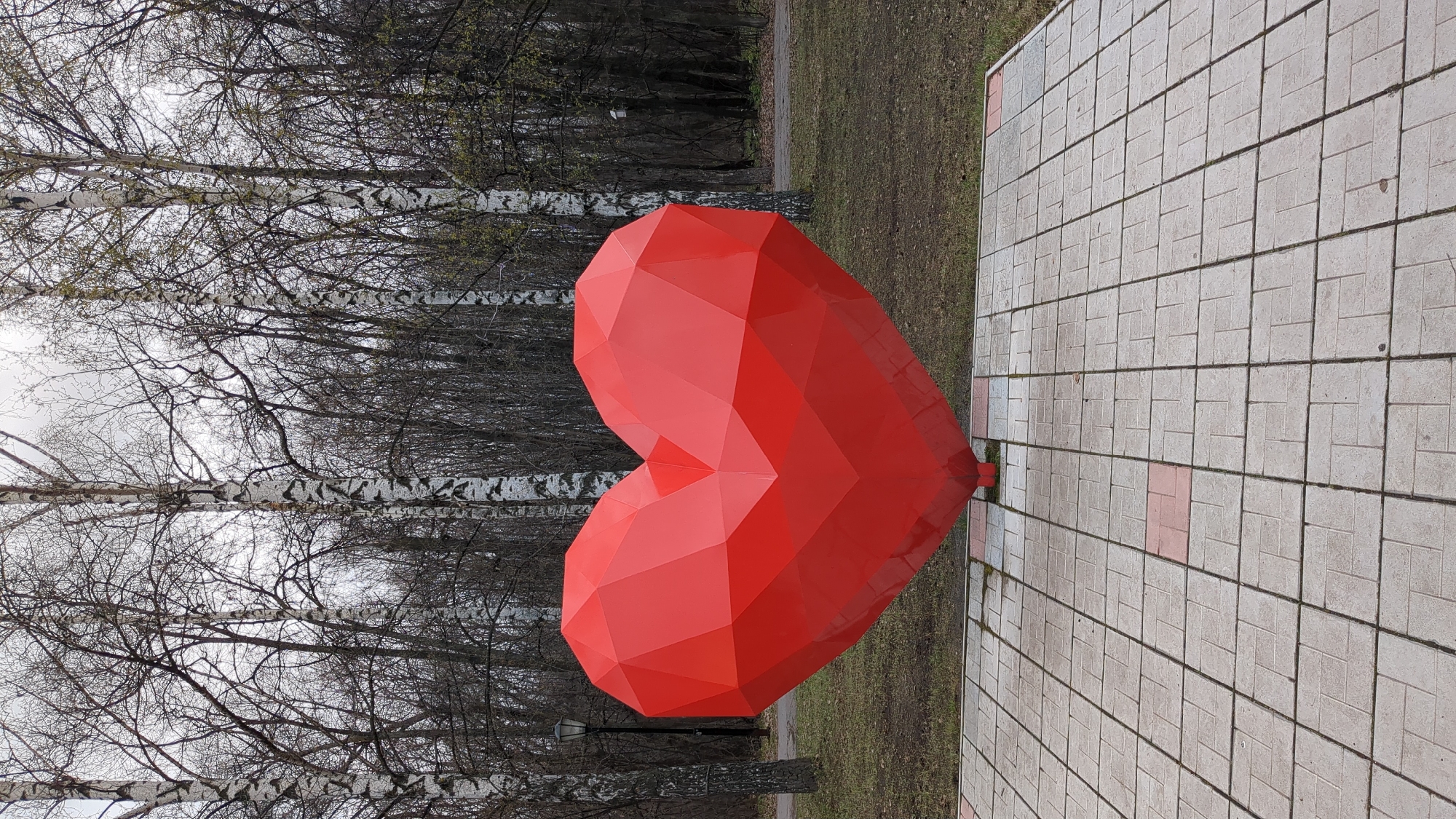 A low-angle, outdoor, medium shot captures a bright red, polygonal heart sculpture. The sculpture is situated on a square-tiled walkway, adjacent to a grassy area with a bare, leafless birch tree forest in the background. The heart sculpture is oriented vertically. The walkway consists of gray square tiles with a few isolated red tiles. The trees in the forest are mostly bare, with some budding leaves. The sky is overcast. The photo was taken in the city of Ulyanovsk, Russia.