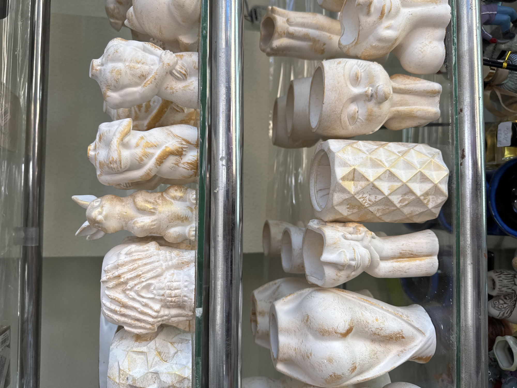 The image displays a vertical arrangement of decorative ceramic or plaster objects, likely small planters or holders, presented in a retail setting. The items are predominantly white with distressed gold accents, creating a weathered or antique look.
The subjects on display are varied. On the left side, objects resembling animal heads, specifically pigs and bunnies with distinct ears and snouts, are visible. Below them, there are items with textured surfaces, one appearing like an abstract human form with horizontal striations.
On the right side of the display, separated by a reflective glass panel and a chrome pole, more objects are seen. These include abstract human-like faces with closed eyes, and a cylindrical object with a prominent geometric, possibly pineapple-inspired, pattern. Further down, another object resembles a stylized human figure or bust. Many of these items feature an open end, suggesting they are designed to hold something.
The scene depicts a static shop display, with the objects meticulously arranged. Reflections in the glass and chrome surfaces provide glimpses of more identical items and the immediate surroundings. In the blurred background on the far right, a small blue pot and a tiny figurine of a person in a purple top and dark pants can be partially discerned. A small white price tag is barely visible, with the number "100" and possibly other characters faintly legible, likely indicating a price. No specific time of day or weather is evident, as it appears to be an indoor or sheltered display. The location is specified as Sochi, Russia, though no explicit visual cues in the image confirm this.