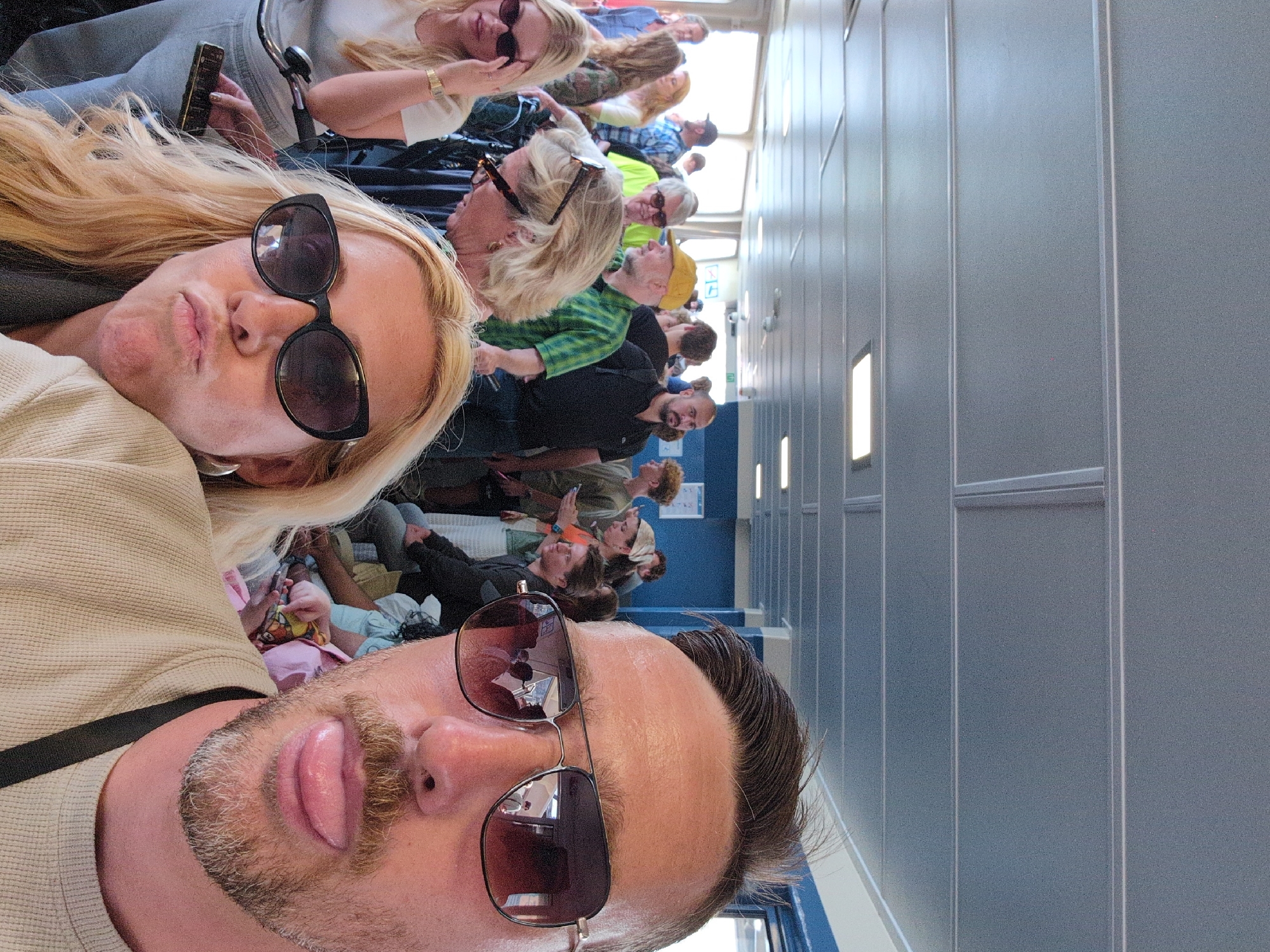 The image shows a group of people inside a ferry, likely in the city of Amsterdam, Netherlands. The ferry appears to be crowded, with people standing and sitting. In the foreground, a man is taking a selfie, sticking his tongue out, and wearing aviator sunglasses. Next to him, a woman is also posing for the selfie, puckering her lips and wearing large black sunglasses. The background is filled with other passengers, some looking towards the camera, others engaged in conversation or looking at their phones. The interior of the ferry has blue walls and a grey ceiling with fluorescent lighting. The overall atmosphere suggests a casual and lively ferry ride.