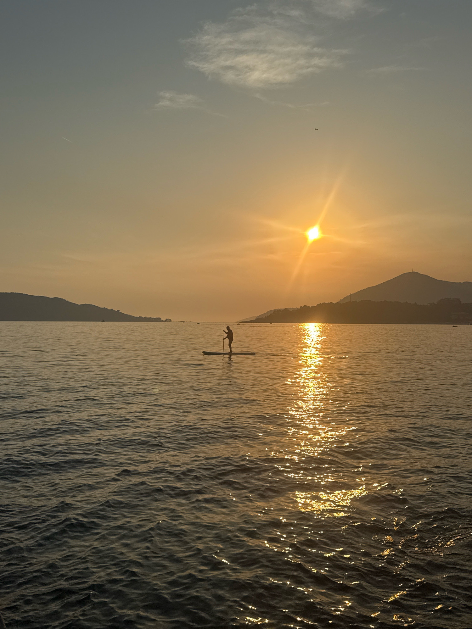 A lone individual stands on a paddleboard in the Adriatic Sea, silhouetted against a vibrant sunset over the mountains of Rafailovići, Montenegro. The sun, a bright orb with prominent rays, is positioned slightly left of center in the frame, casting a shimmering golden path across the rippling water. The water itself is a deep, dark blue, reflecting the warm hues of the sky. In the background, the rugged coastline of Rafailovići is visible, its silhouette softened by the fading light. Wisps of clouds drift across the expansive sky, adding texture to the scene. A faint contrail from an aircraft can be seen high in the sky, and a small dark shape, possibly a bird, is visible to the right of the sun. The overall atmosphere is one of tranquility and natural beauty.