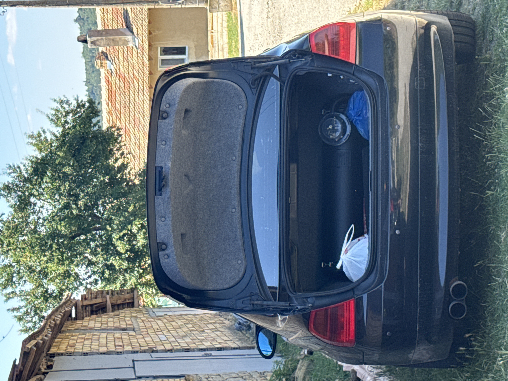 The image shows the open trunk of a black car parked on a grassy area. Inside the trunk, there is a gas cylinder, a white plastic bag, and a blue plastic bag. In the background, there is a brick building on the left, and a house with a tiled roof and a chimney on the right. There are trees and hills in the distance, under a clear blue sky. The scene is set in the city of Konop, Bulgaria.