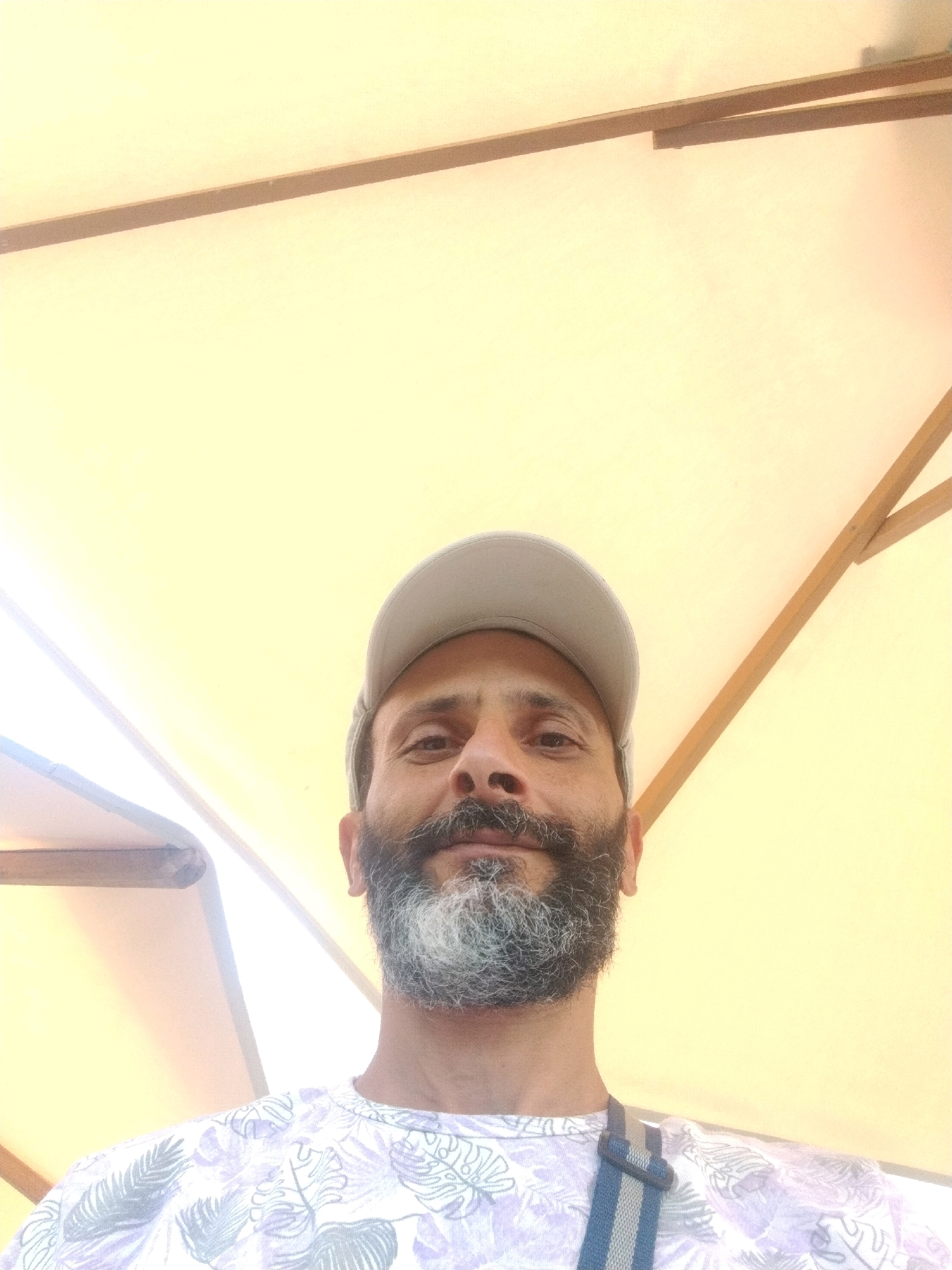 A man with a full beard and a gray baseball cap is looking up at the camera. He is wearing a light-colored t-shirt with a floral pattern. The background is a pale yellow umbrella or canopy, with wooden supports visible at the top. The image appears to be taken outdoors, possibly in Monastir, Tunisia, given the information provided.