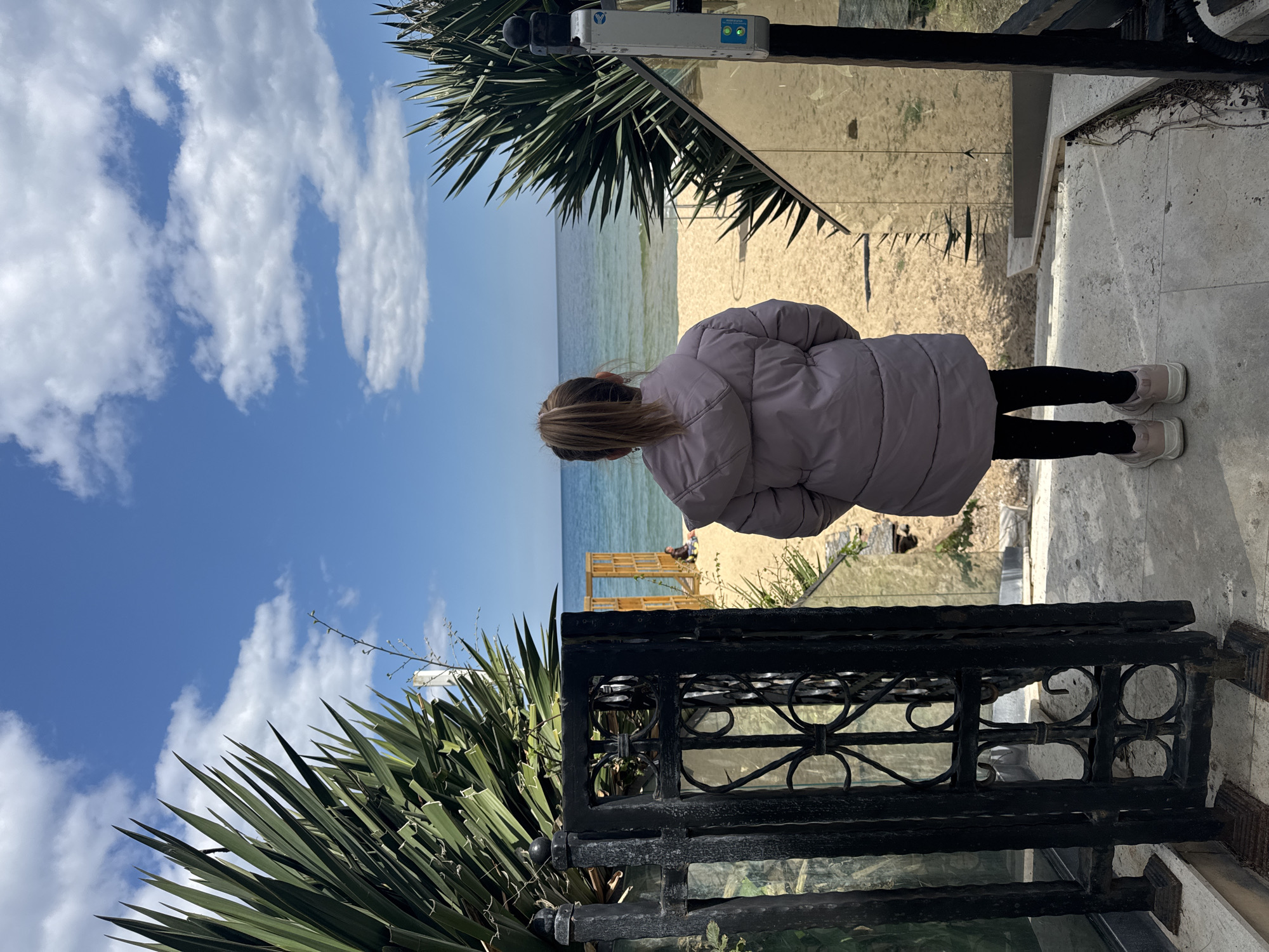 A person stands on a ledge overlooking a beach. The person, seen from the back, wears a light purple puffer jacket, black leggings, and light-colored sneakers. The beach has golden sand and blue water. A metal railing and leafy plants are in the foreground, and the sky is blue with white clouds. The scene likely takes place in Varna, Bulgaria, due to its coastal location and architectural elements, although specific landmarks are not visible to confirm the precise location within the city.