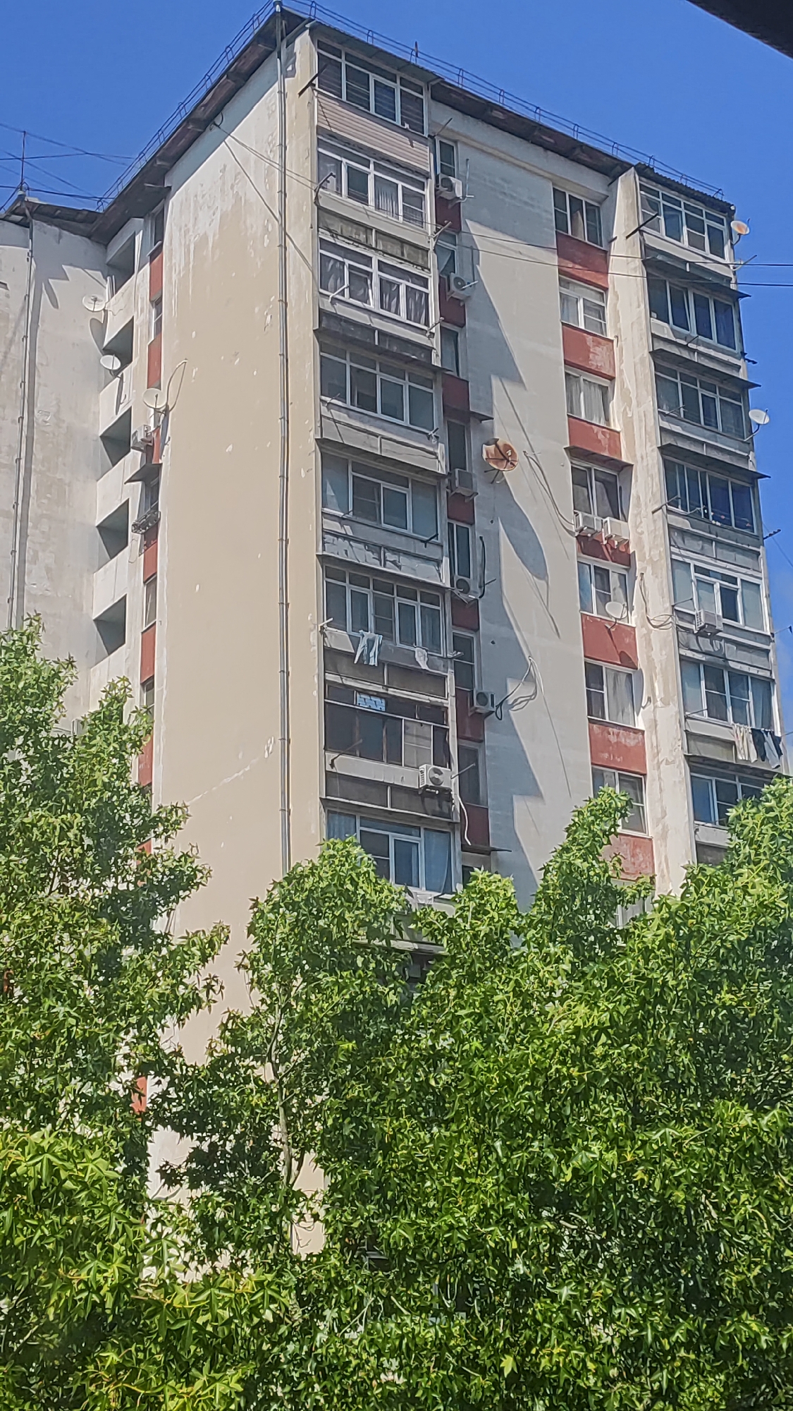 A multi-story apartment building is visible in Dagomys, Russia, against a clear blue sky. The building features a light-colored facade with red accents around some of the windows and balconies. Numerous windows and enclosed balconies are visible, some with air conditioning units mounted outside. Satellite dishes are attached to the exterior of the building. In the foreground, lush green trees partially obscure the lower portion of the building. The scene appears to be a residential area.