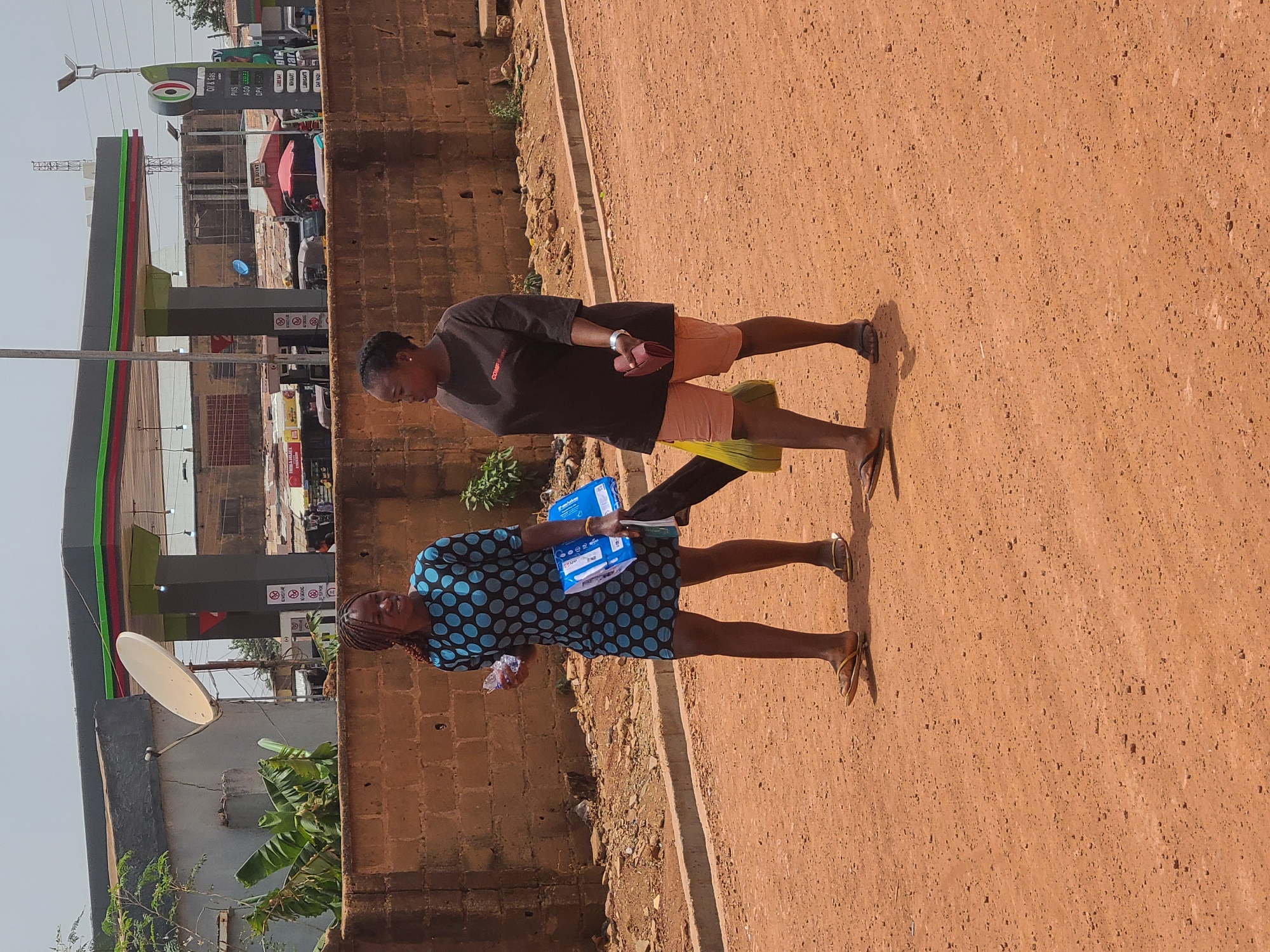 The image shows two women walking on a red dirt road, presumably a sidewalk. The woman in the front is wearing a blue polka dot dress and holding a blue box labeled "Beulahm". The second woman is wearing a black t-shirt and salmon-colored shorts. In the background, a gas station is visible. The surrounding environment includes a brick wall, vegetation, and a satellite dish. Based on the gas station signage "Oil & Gas", it is probably in Jos, Nigeria.