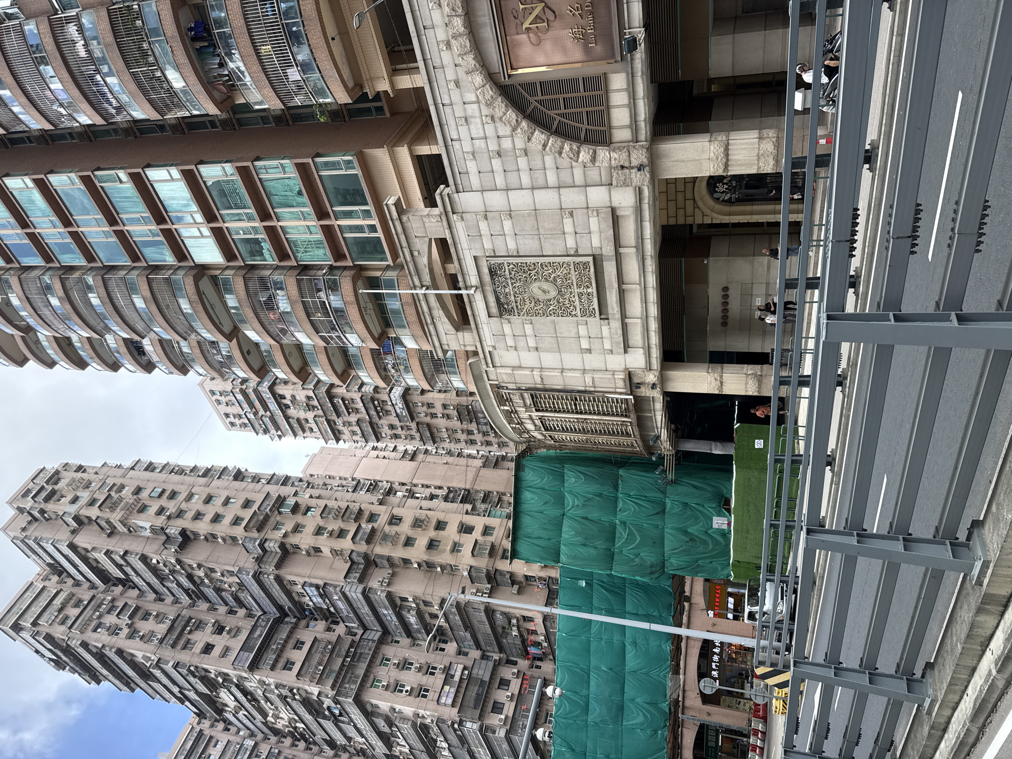 This is an eye-level medium shot of a street scene in Macau, China, with several tall buildings in the background. The foreground features a grey metal barrier and a road with white lane markings. On the road, there are a few vehicles, including a white truck. To the left of the road, there is a raised sidewalk with a metal fence. A pedestrian bridge with a grey metal railing is visible across the street.
In the midground, the scene is dominated by a large, modern building with a curved facade and multiple balconies with glass railings. The building is adorned with decorative stone accents and arched windows. Below this building, there is a construction site covered by green netting. Signs in Chinese characters are visible on some of the storefronts, suggesting a commercial area.
The background reveals several very tall, older apartment buildings with numerous balconies, air conditioning units, and windows. The architecture of these buildings is dense and somewhat utilitarian. The sky is overcast. A few people are visible on the sidewalks and near the entrance of one of the buildings. The overall impression is of a bustling urban environment in Macau.