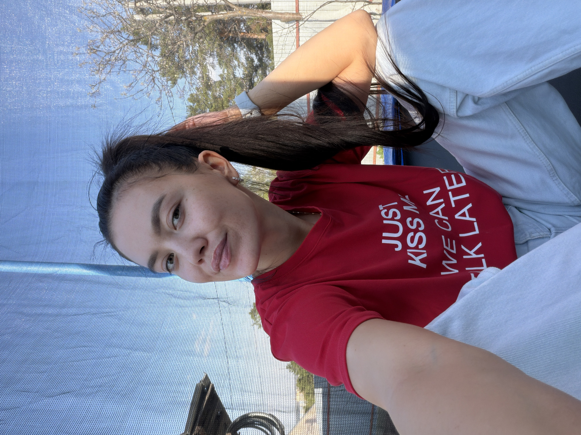 A young woman with dark hair pulled back in a ponytail is the subject of this selfie. She is wearing a red t-shirt with white text that reads "JUST KISS ME, WE CAN TALK LATER" and light-colored jeans. She appears to be outdoors, sitting on a trampoline with a blue mesh safety net. The background includes trees, a fence, and what appears to be part of a building. Based on the location provided, the scene is situated in the city of Chaikovsky, Russia. The woman is holding the camera, taking a picture of herself with a slight smile.