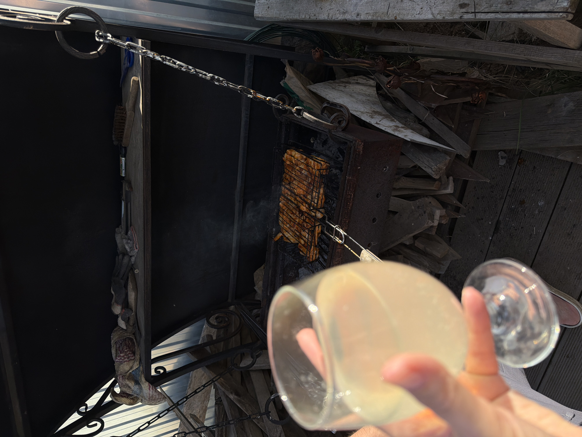 The image shows a person holding a glass with a light-colored liquid in front of a barbecue grill. The grill has food cooking on it and is suspended by a chain. There is a pile of wood next to the grill. Gloves, a brush, and other objects are hanging on the barbecue rack. The environment suggests an outdoor setting, possibly a backyard or recreational area. The location is in the city of Chaikovsky, Russia.