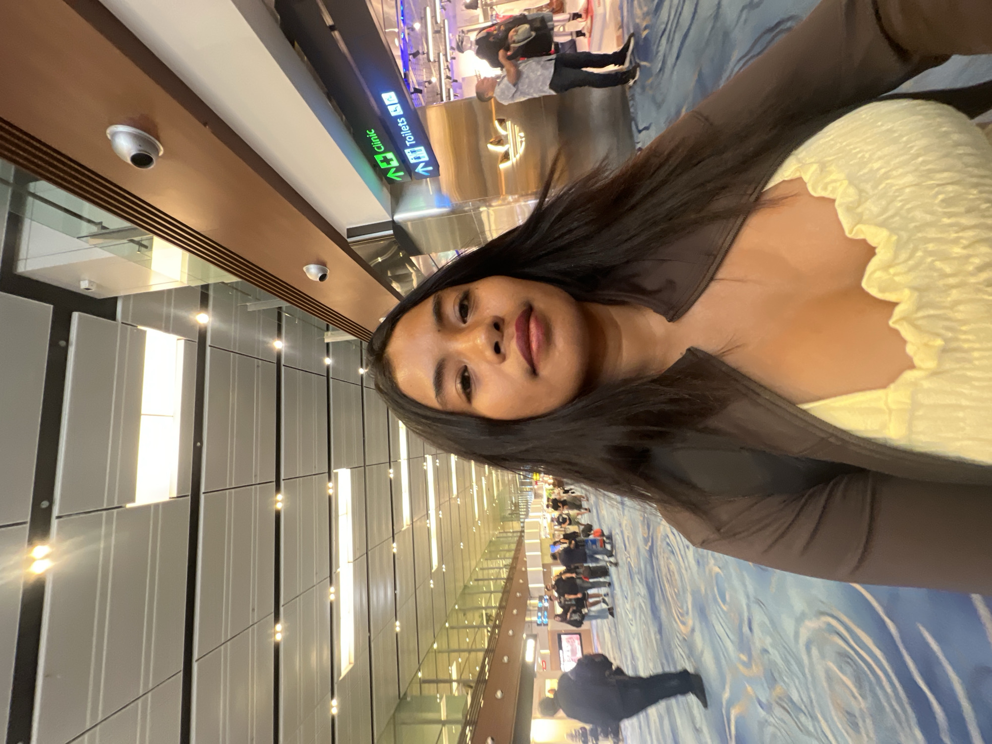 A young woman with long, dark hair is taking a selfie in a brightly lit airport terminal. She is wearing a brown jacket over a yellow, ruched top. In the background, other travelers are walking through the terminal, some with luggage. Signs indicate "Clinic" and "Toilets". The floor has a blue and white patterned carpet. The environment appears to be an indoor public space with modern architecture, consistent with an airport.