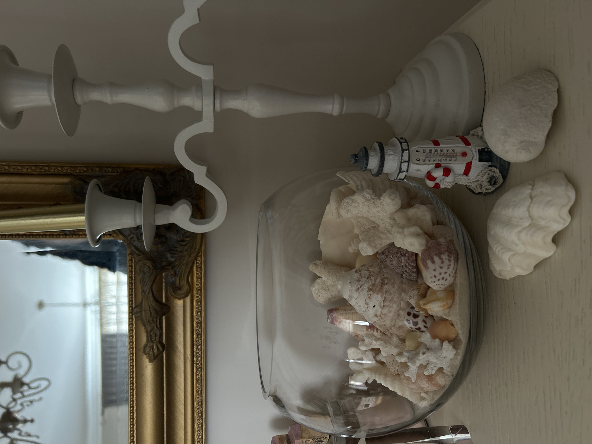The image depicts a still life arrangement with a nautical theme. A clear glass bowl filled with shells and coral is prominently displayed. Next to the bowl is a decorative lighthouse thermometer. Two white seashells rest on the surface beside the lighthouse. In the background, a white candlestick holder and a gold-framed mirror are visible. The scene appears to be set on a light-colored shelf. The location in Plovdiv, Bulgaria, cannot be determined from the image.