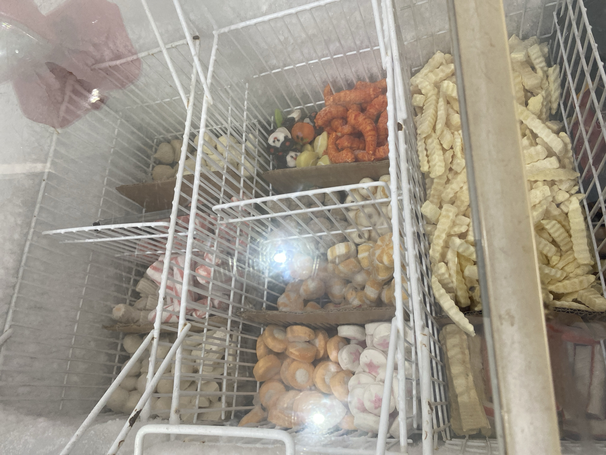 A freezer case filled with various frozen food items is shown. The freezer is divided into multiple shelves, each holding different products. On the left side, crinkle-cut french fries are visible. The middle shelves contain an assortment of circular processed food items, some white with pink accents, others orange, and some appearing to be small, round meatballs or fish balls. There are also some novelty-shaped items, possibly for hot pot, including an orange pumpkin-shaped item and some black and white cat-shaped items. On the right, more white spherical items are stacked. A pink, possibly fabric or plastic item, is visible on the bottom right. The freezer interior is covered in frost. The image is taken from a slightly angled perspective, looking into the freezer through a glass door. The location is identified as Payatieng, Indonesia.