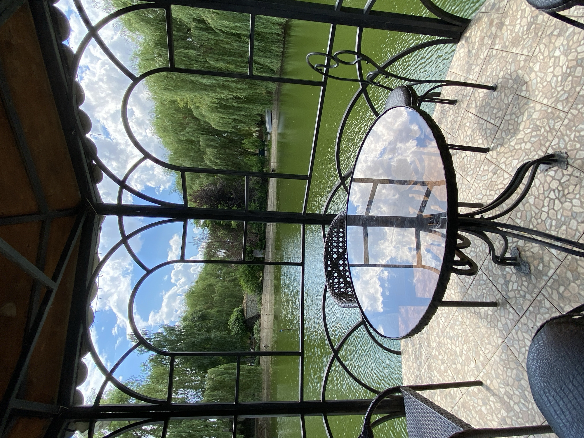 A glass-topped table with a metal base sits on a patterned tile patio. The table is reflecting the sky, which is filled with white, fluffy clouds. Behind the table, a body of green water is visible, likely a pond or lake, surrounded by lush green trees. The scene appears to be set in an outdoor gazebos or covered patio area, with a decorative metal railing. The time of day appears to be daytime, with bright sunlight casting shadows. The weather seems pleasant, with a clear sky and evident clouds. There are no people or text visible in the image. The overall impression is one of a peaceful, natural setting, possibly a restaurant or cafe with an outdoor seating area overlooking a scenic view.