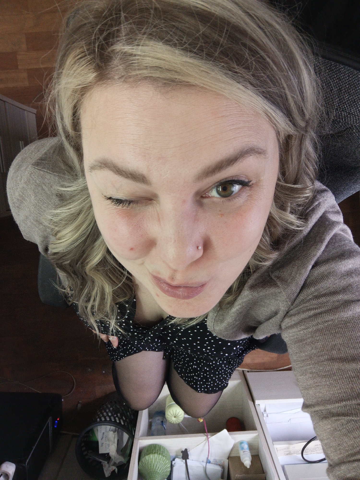 A woman with blonde hair is looking upwards at the camera with a wink. She has a nose piercing, a mole on her left cheek, and is wearing a black dress with white polka dots and a light brown cardigan. She is positioned above an open drawer containing various small items including yarn, a bottle, and other supplies. Her legs, clad in black tights, are visible between her dress and the drawer. A black object with a blue light is in the background to the left, and a trash bin with contents is also visible. The floor is a reddish-brown color. There is no information in the image that indicates this is in the city of Noviy, Russia.