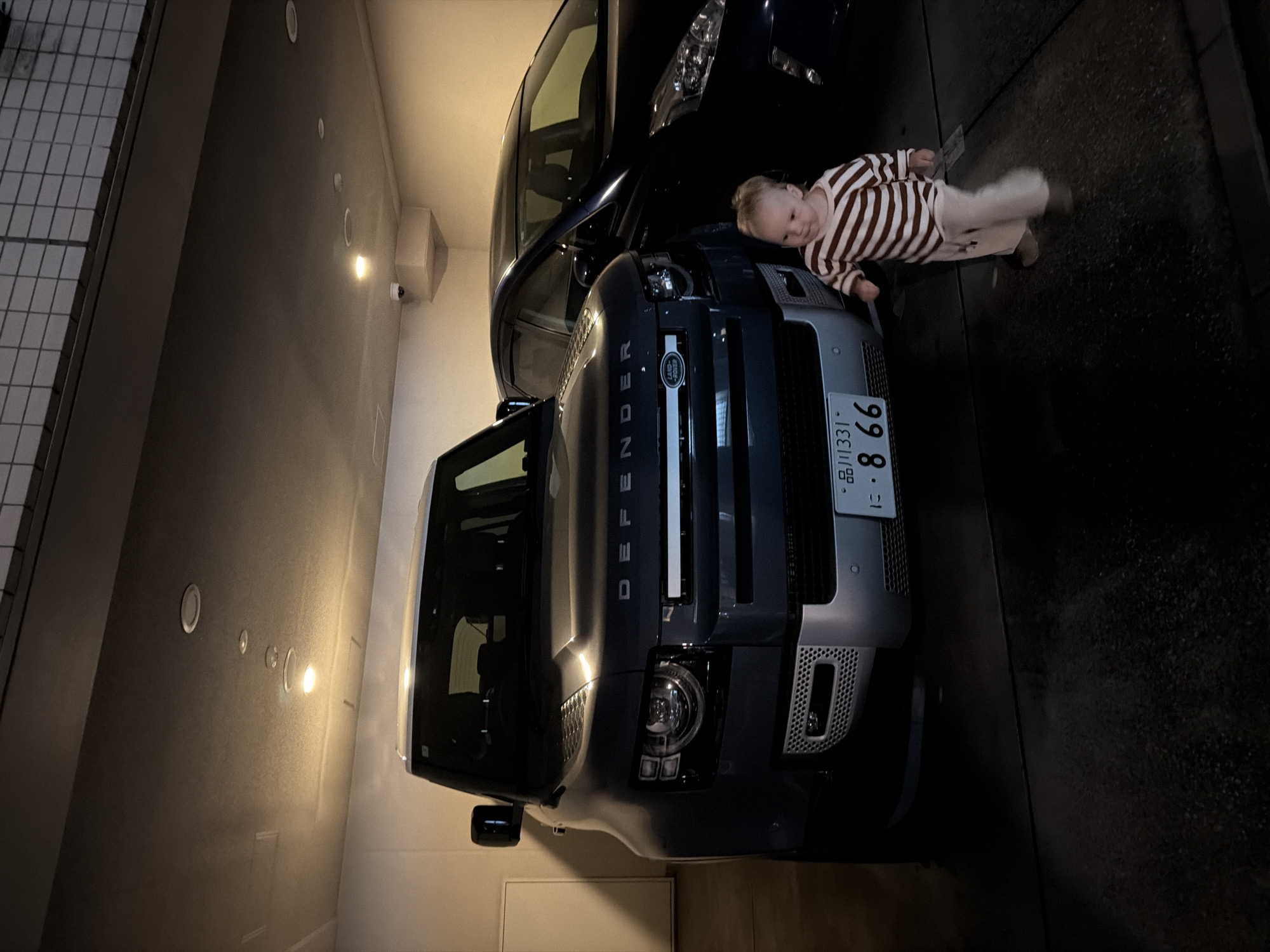 In the city of Chiyoda, Japan, within a dimly lit, covered parking structure, a dark blue Land Rover Defender is parked. The vehicle's front is facing the viewer, with its headlights illuminated. To the right of the Defender, a dark-colored sedan is partially visible. In the foreground, a young child, appearing to be a toddler, is walking towards the viewer. The child is wearing a striped long-sleeved shirt and light-colored pants. The license plate of the Defender is visible and reads "品川331" and "・866". The environment appears to be an indoor parking garage, with exposed ceiling lights and concrete walls.