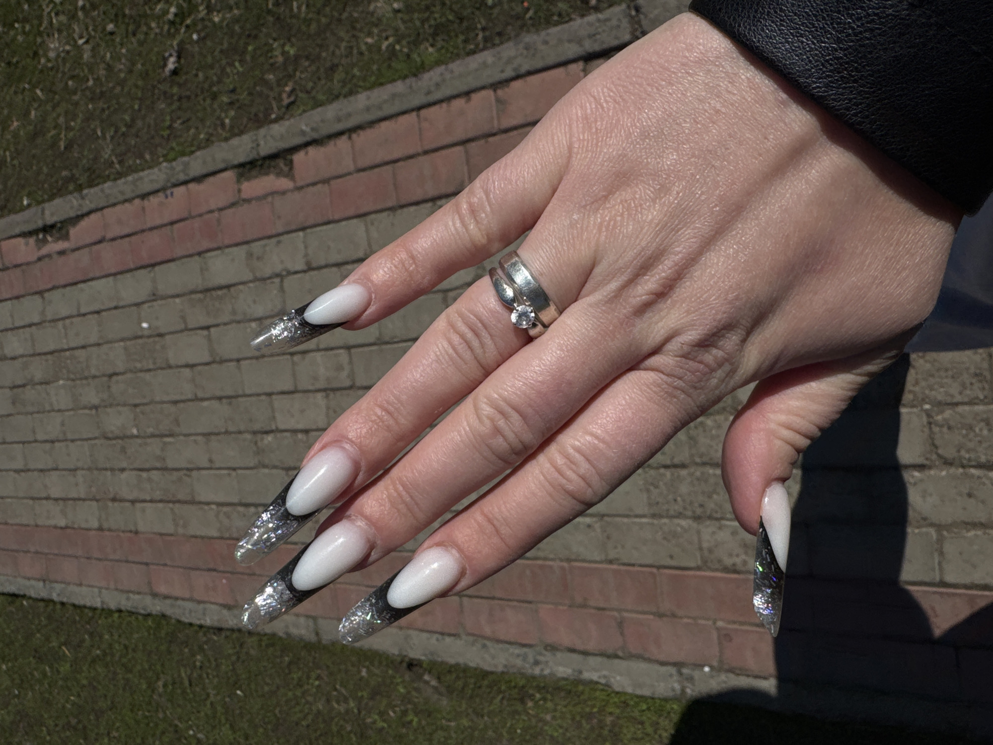 A close-up shot displays a female hand adorned with long, artificial nails with white tips and sparkly black accents. The hand wears a silver wedding band set, with a solitaire diamond on the engagement ring. The hand is positioned against a background of a brick walkway with patches of green grass alongside it. A small portion of a black leather jacket sleeve is visible in the upper right corner. The location is an outdoor area in a city.