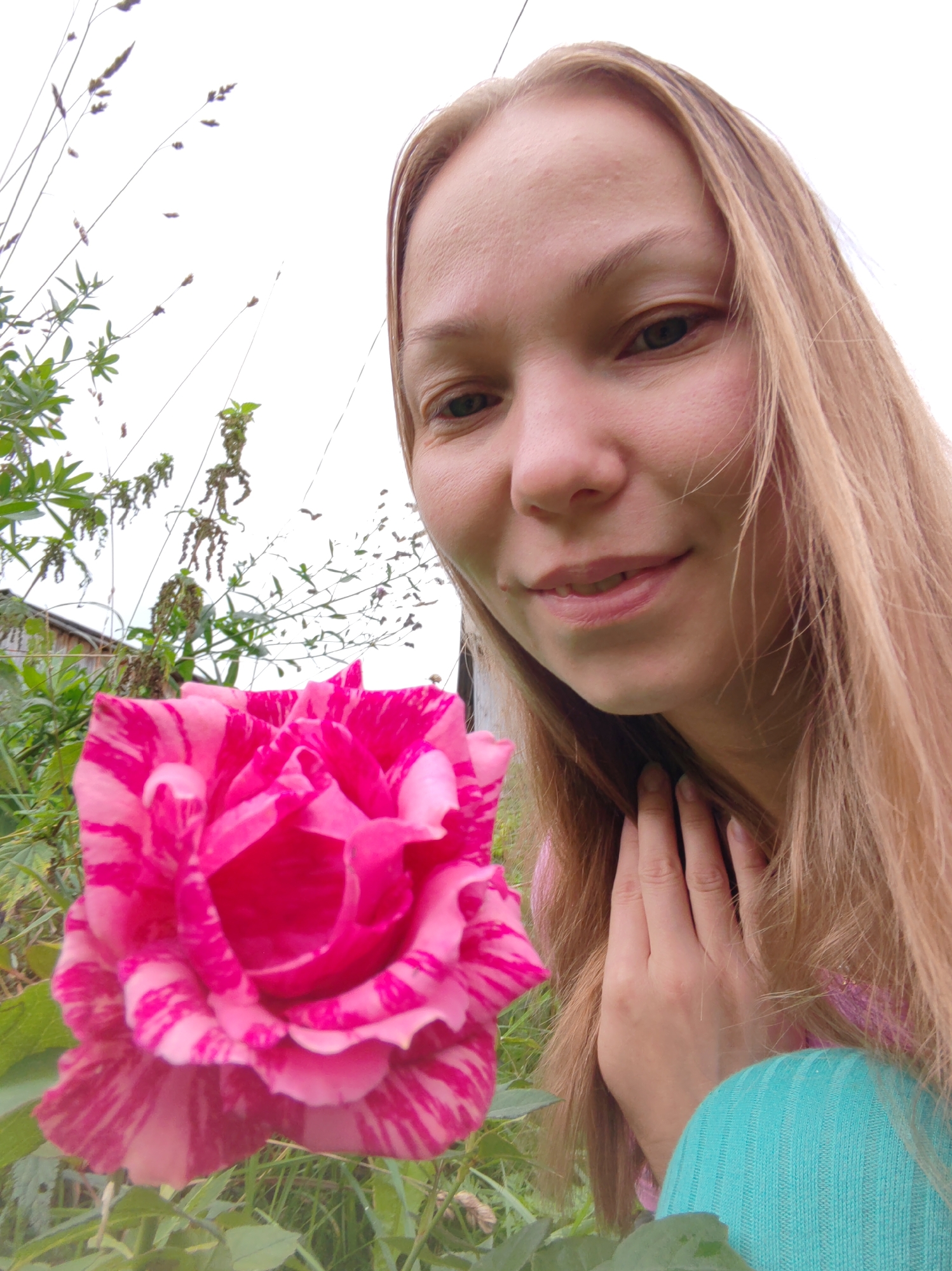 A young woman with long blonde hair is in a garden with a pink and white striped rose in the foreground. She is holding her hand to her face, and her body is partially visible on the right side of the frame, wearing a turquoise top. The background shows greenery and some dried plants. The image appears to be a selfie, taken outdoors. There is no specific information that can identify the location as Чухлэм, Russia.